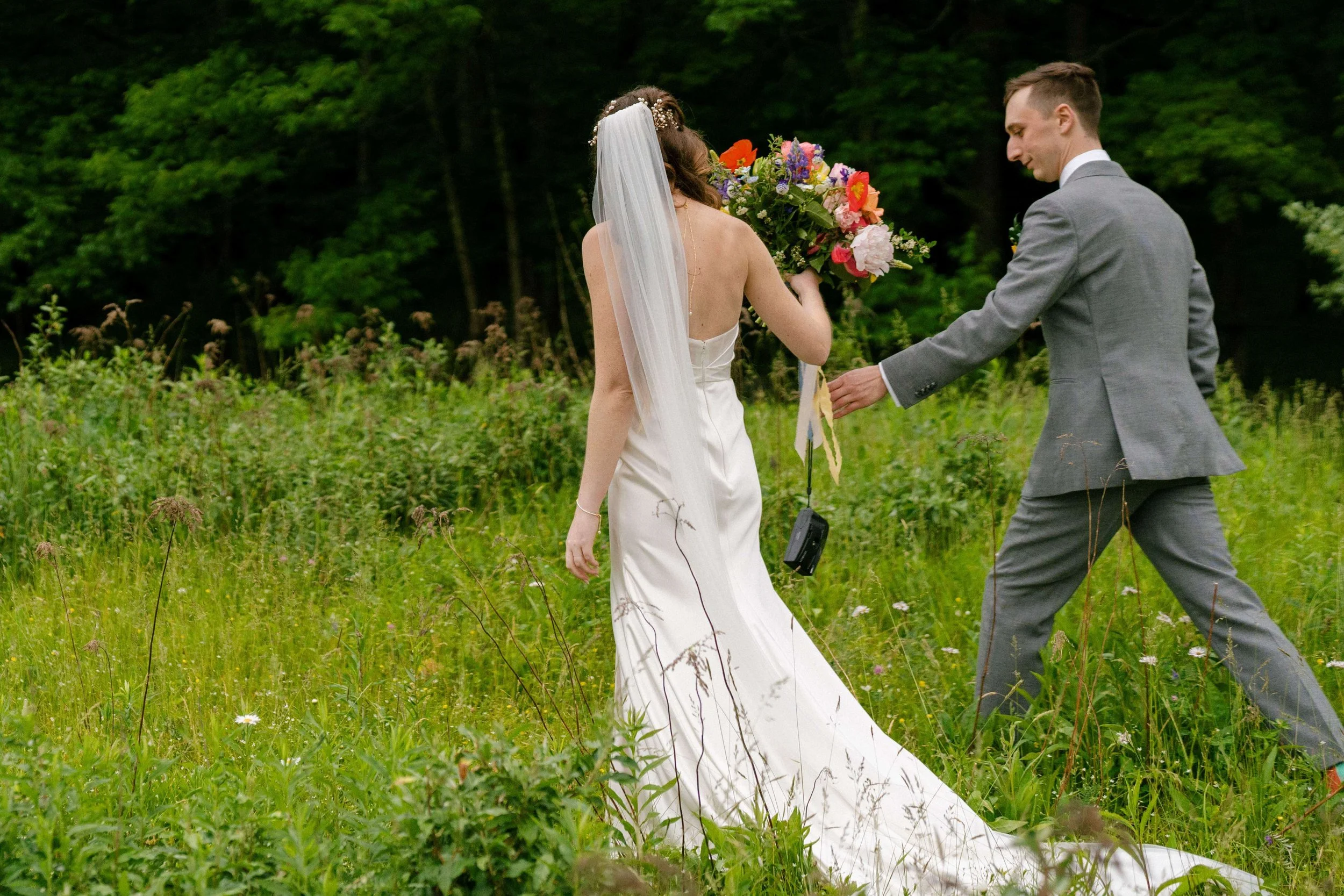 Bride and groom walking through a lush New England wildflower meadow, groom carrying a vibrant bouquet of orange poppies, pink peonies, and purple wildflowers with ribbon streamers, Milkweed Floral Co