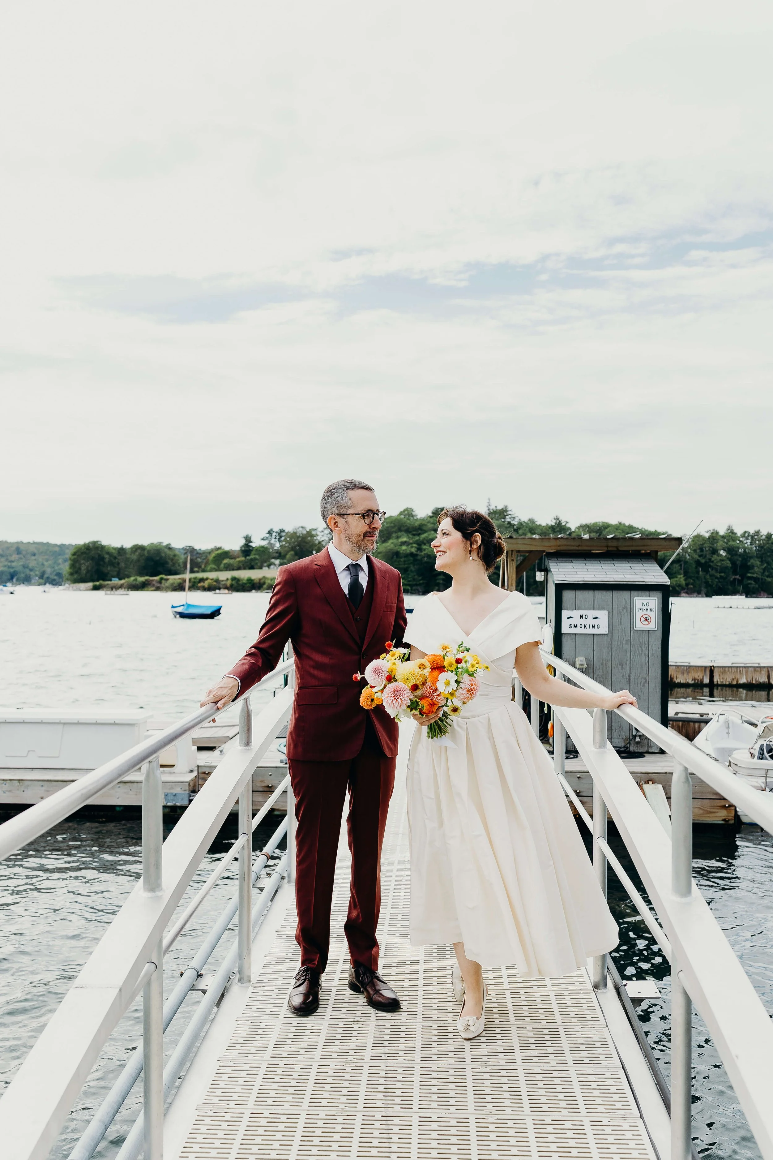 Bride and groom on a coastal Maine pier, bride holding a vibrant garden bouquet of dahlias, zinnias, and cosmos by Milkweed Floral Co