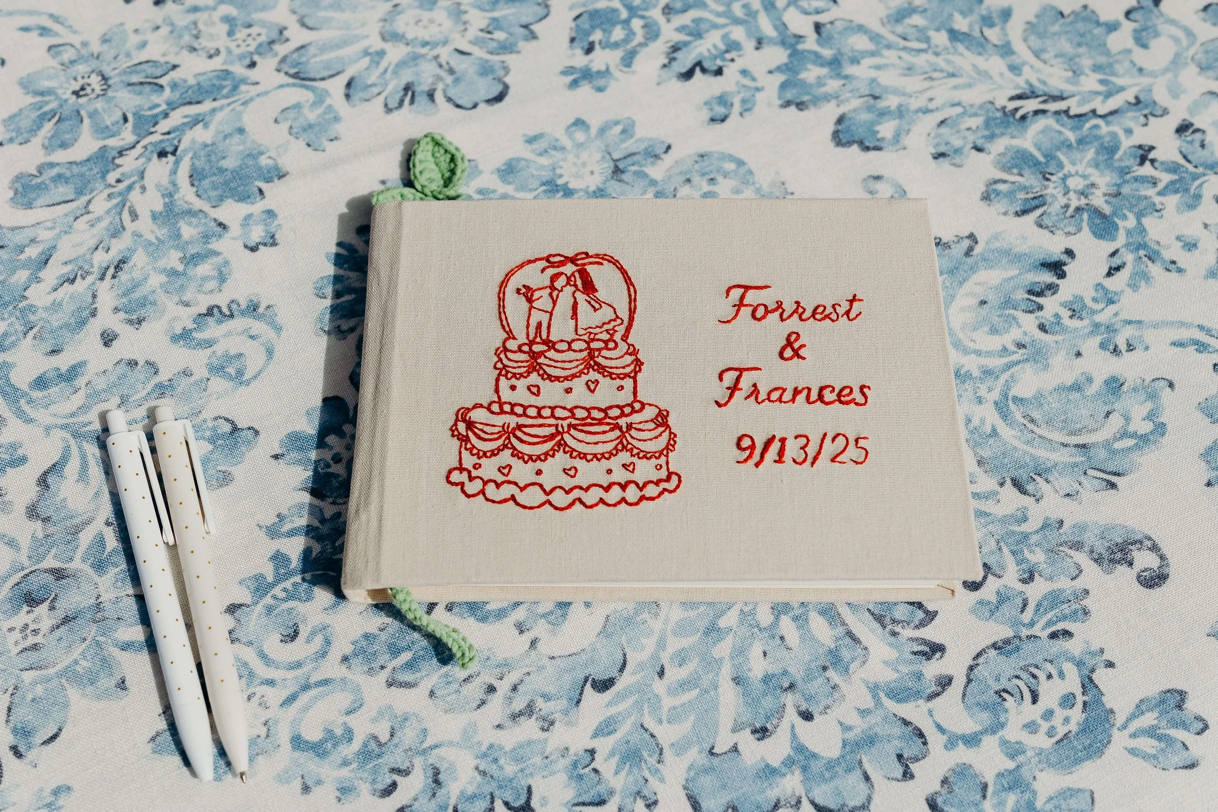 Personalized linen guest book embroidered with a wedding cake and "Forrest & Frances 9/13/25" on a blue floral tablecloth, Maine wedding by Milkweed Floral Co