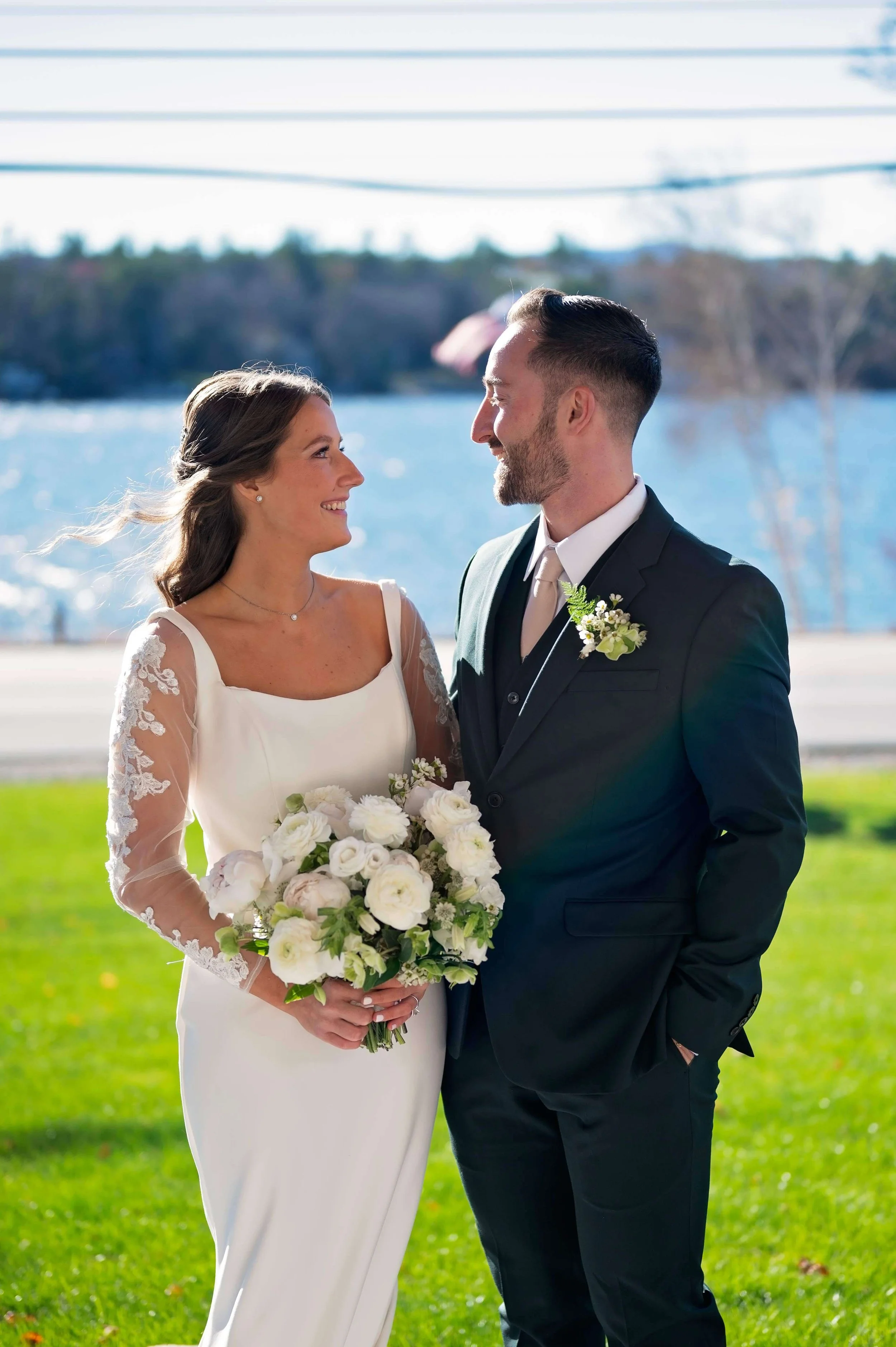Bride and groom smiling at each other on the waterfront lawn, bride holding a white peony and ranunculus bouquet, groom wearing a waxflower and fern boutonniere at Newfound Lake Inn, Bridgewater New Hampshire wedding florals by Milkweed Floral Co