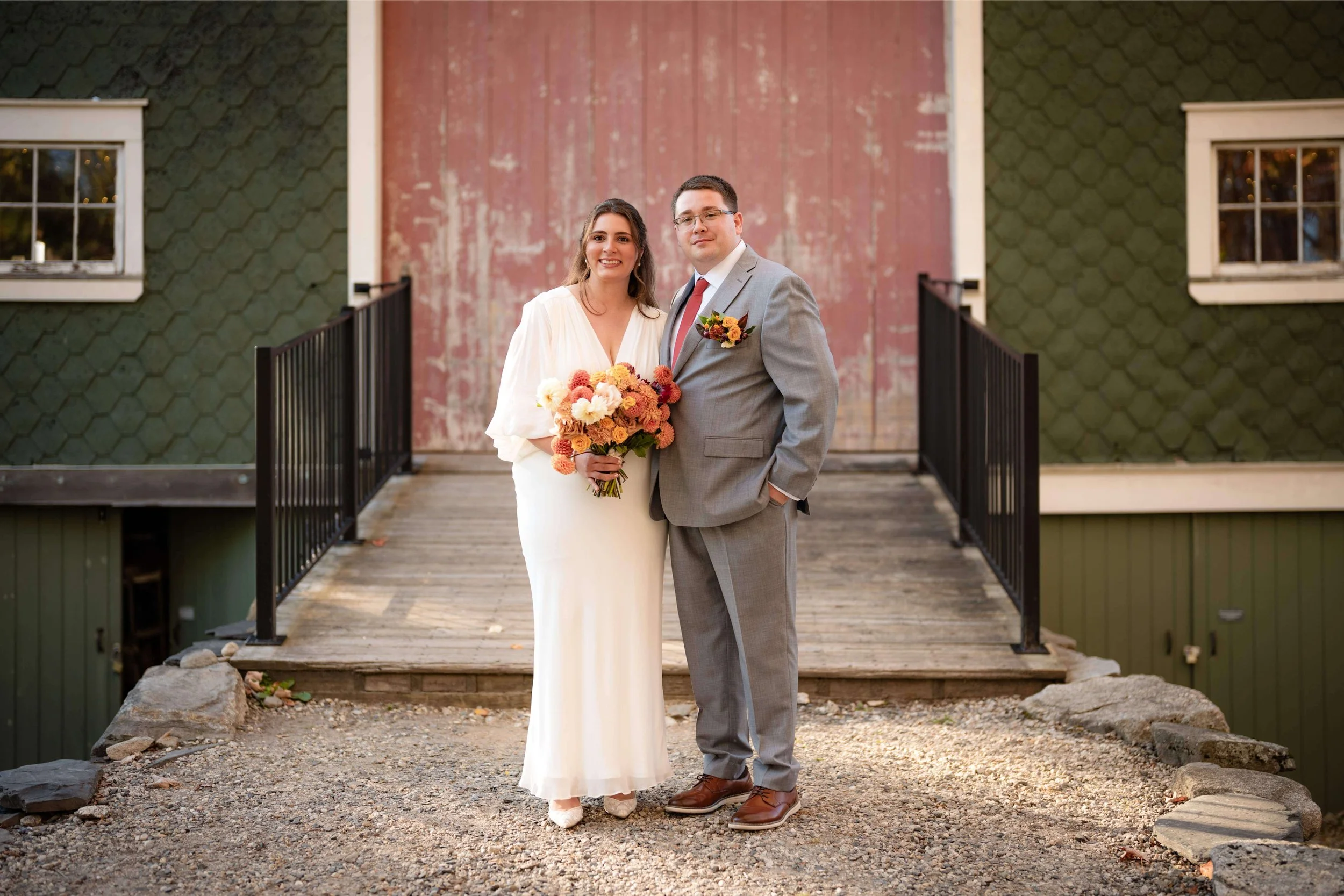 Joyful bride and groom standing in front of red, green, and white barn, bride is holding a bouquet of peach dahlias and yellow roses, groom wears matching boutonniere, Milkweed Floral Co