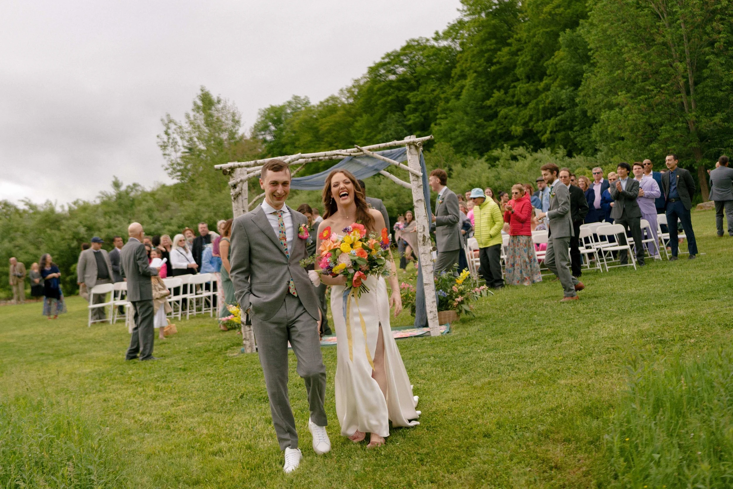 Joyful bride and groom recessional at an outdoor spring wedding with birch arch and vibrant wildflower bouquet, Milkweed Floral Co
