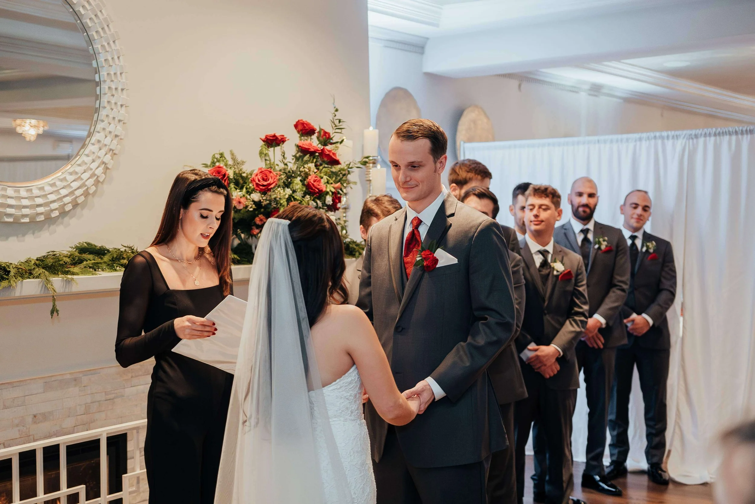 Bride and groom exchanging vows beside a red rose and greenery fireplace arrangement at Saphire Estate, Sharon Massachusetts wedding florals by Milkweed Floral Co
