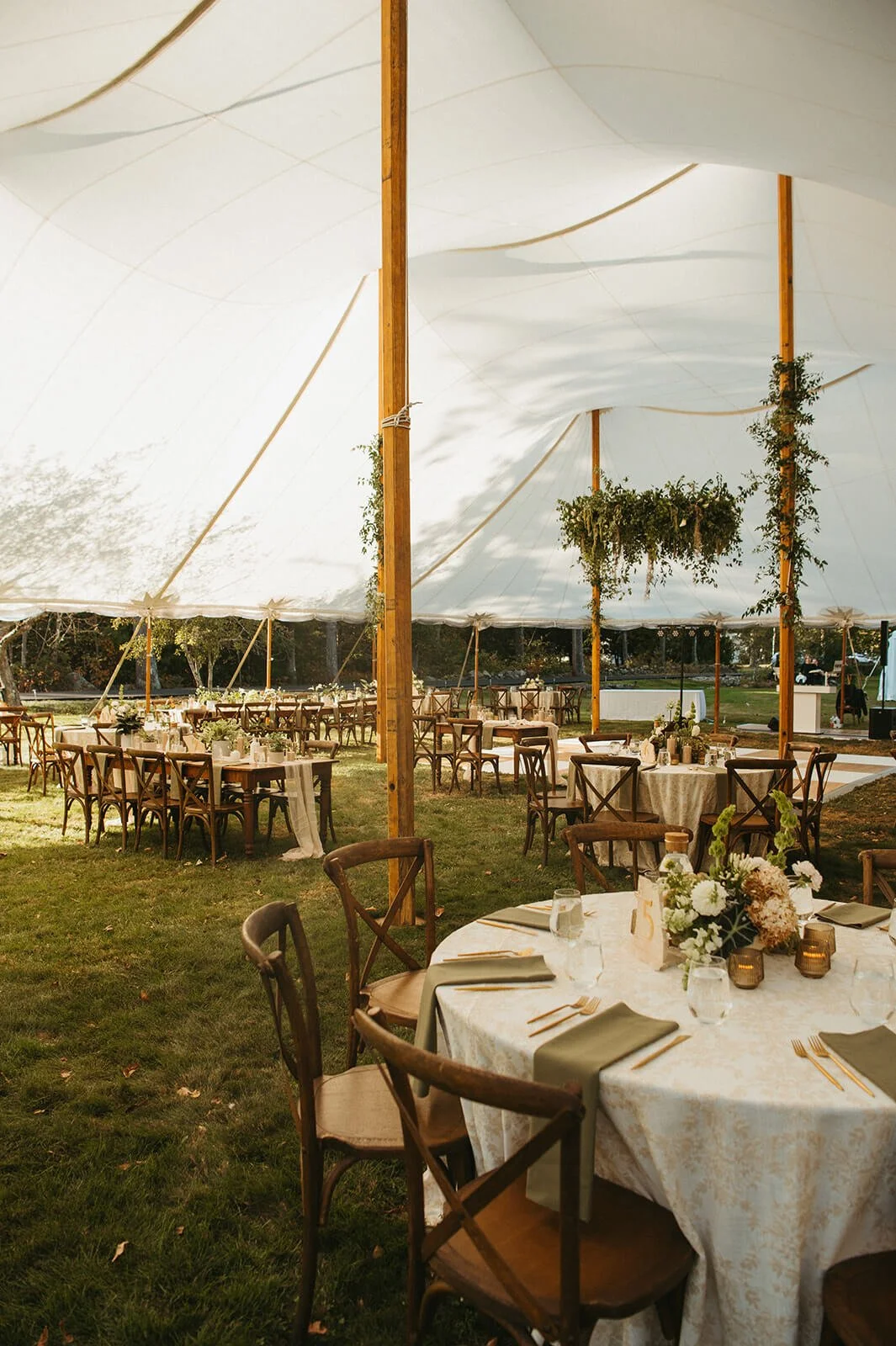 Elegant tented outdoor wedding reception with wood cross-back chairs, sage green napkins, white floral centerpieces, and a greenery-draped tent pole installation, Monson Massachusetts, Milkweed Floral Co
