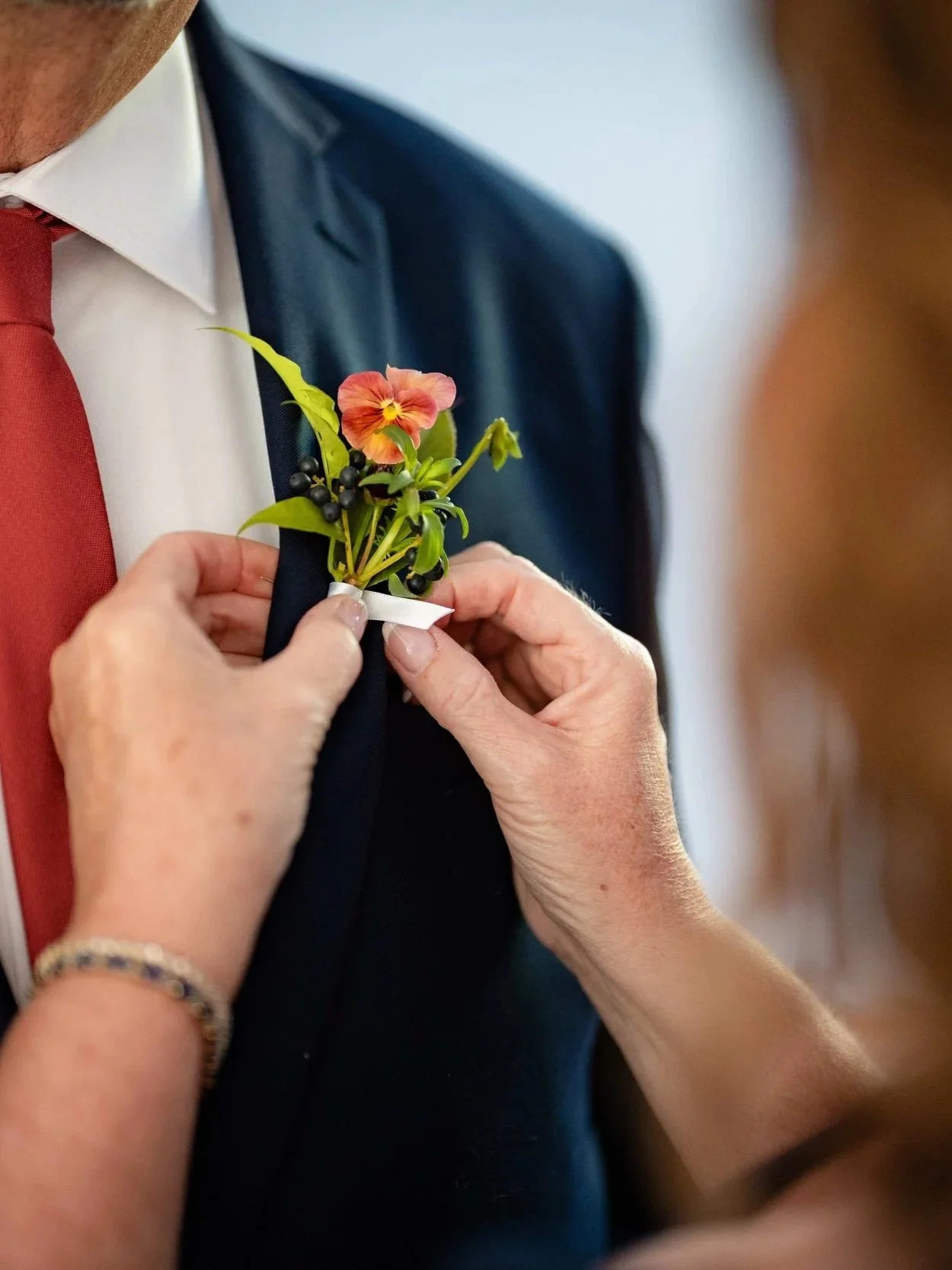 Garden boutonniere with peach pansy and dark berries being pinned to a navy suit, wild-inspired wedding florals by Milkweed Floral Co