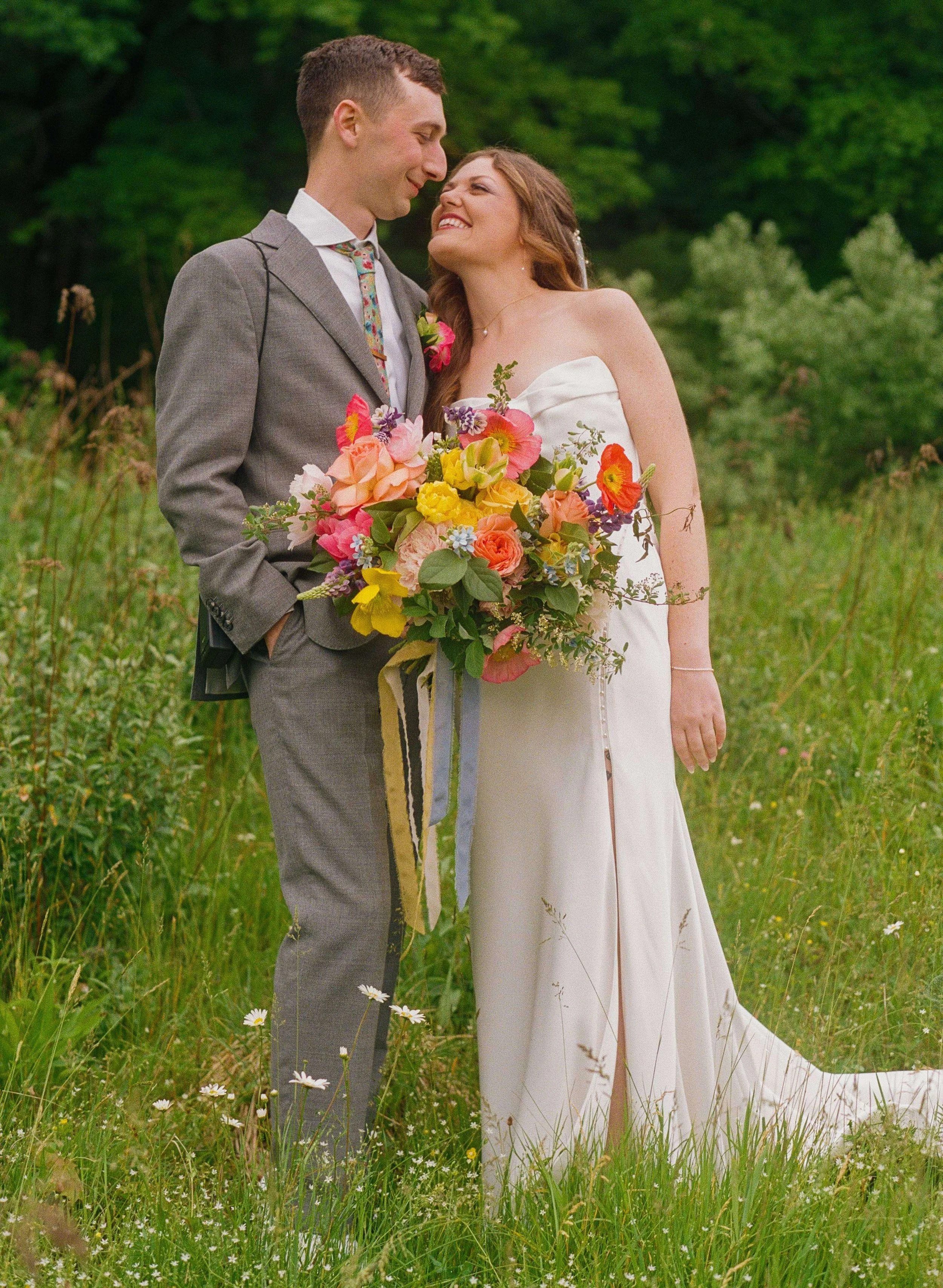 Bride and groom laughing together in a wildflower meadow, bride holding a vibrant bouquet of poppies, garden roses, and yellow tulips with ribbon streamers, New England wedding by Milkweed Floral Co