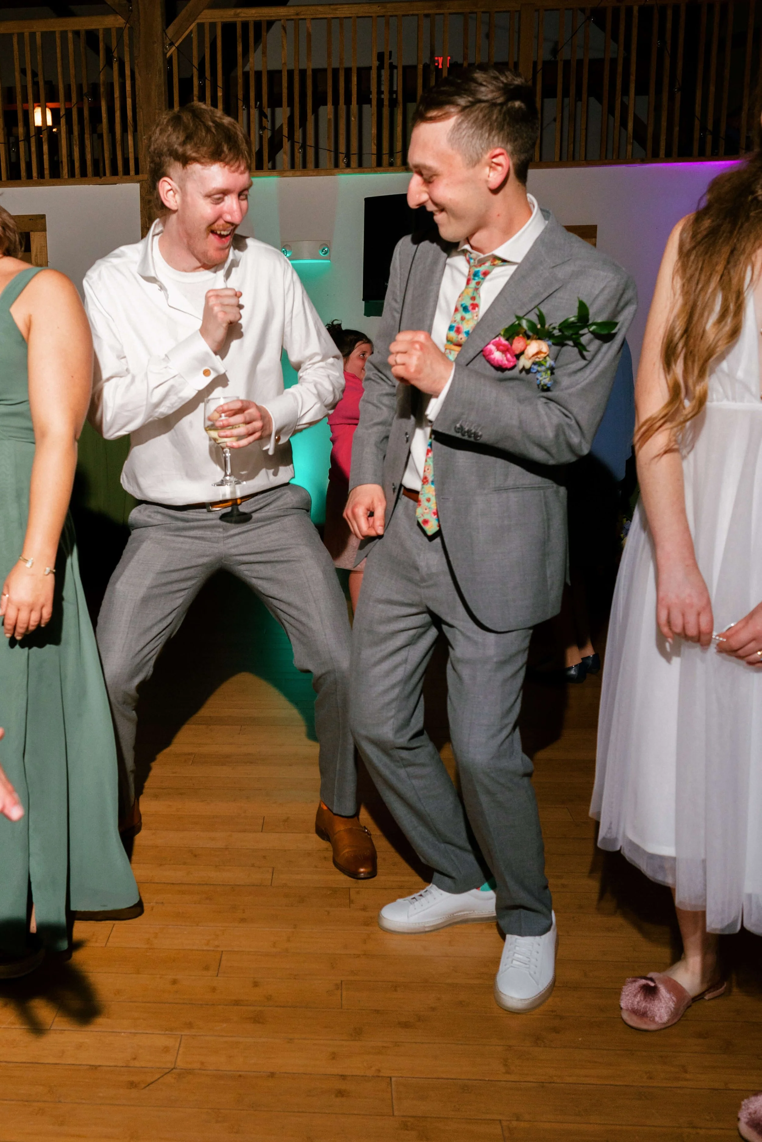 Groom dancing joyfully with a groomsman at an indoor wedding reception, groom wearing a colorful floral boutonniere and floral tie, Montague Retreat Massachusetts, Milkweed Floral Co
