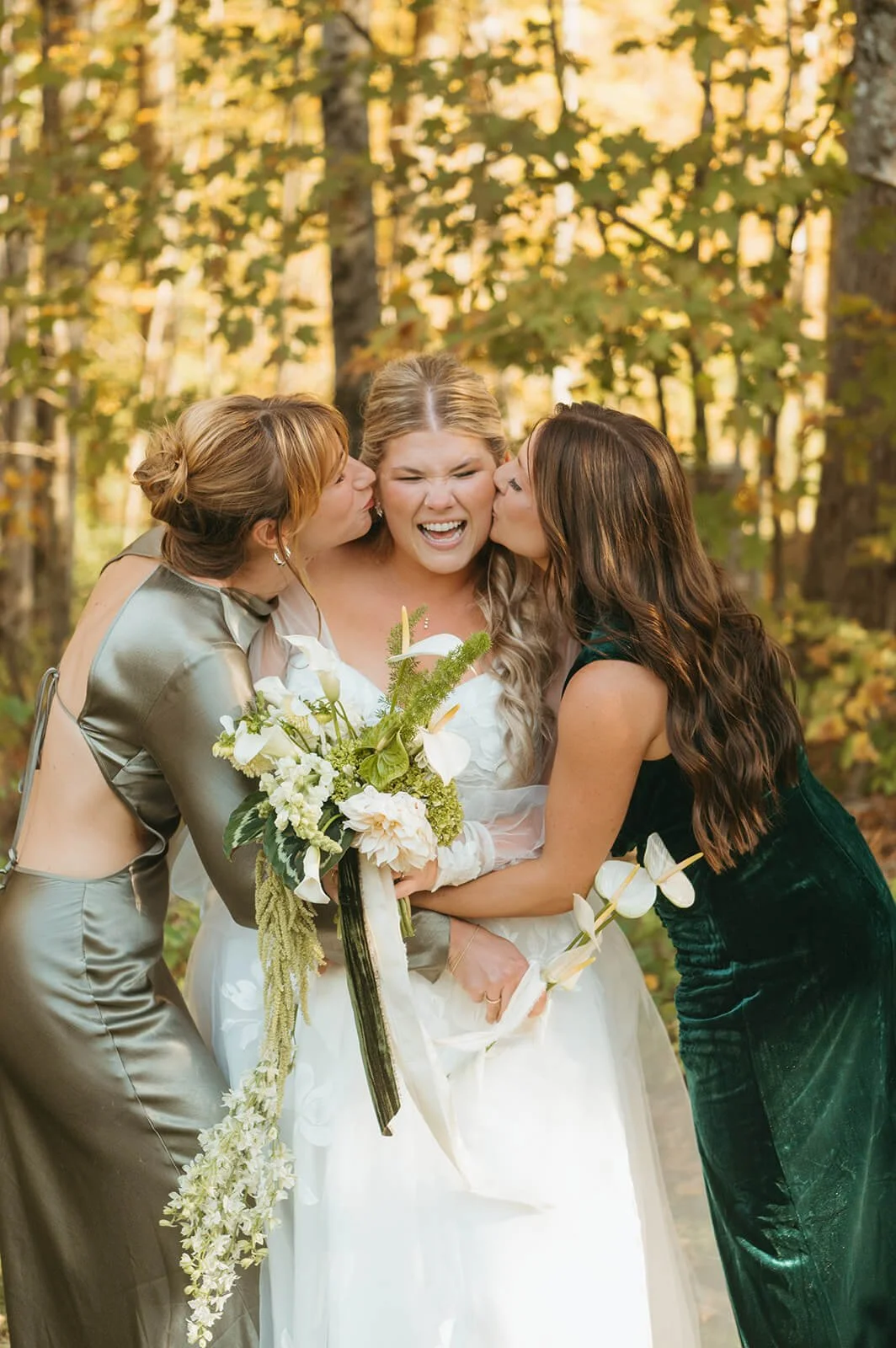 Two bridesmaids in green velvet and satin dresses kissing a laughing bride, bride holding a cascading bouquet of white calla lilies, dahlias, and trailing amaranthus against fall foliage, Milkweed Floral Co