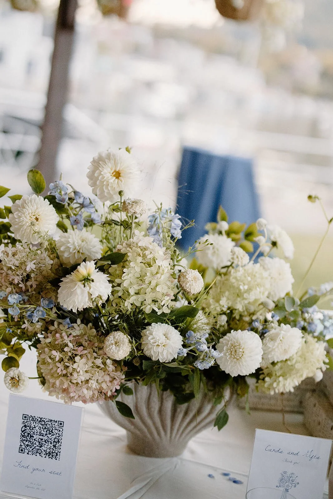 Lush coastal wedding escort card table arrangement with white dahlia pompons, blue delphinium, and blush hydrangea in a textured white urn, Camden Yacht Club Maine, Milkweed Floral Co