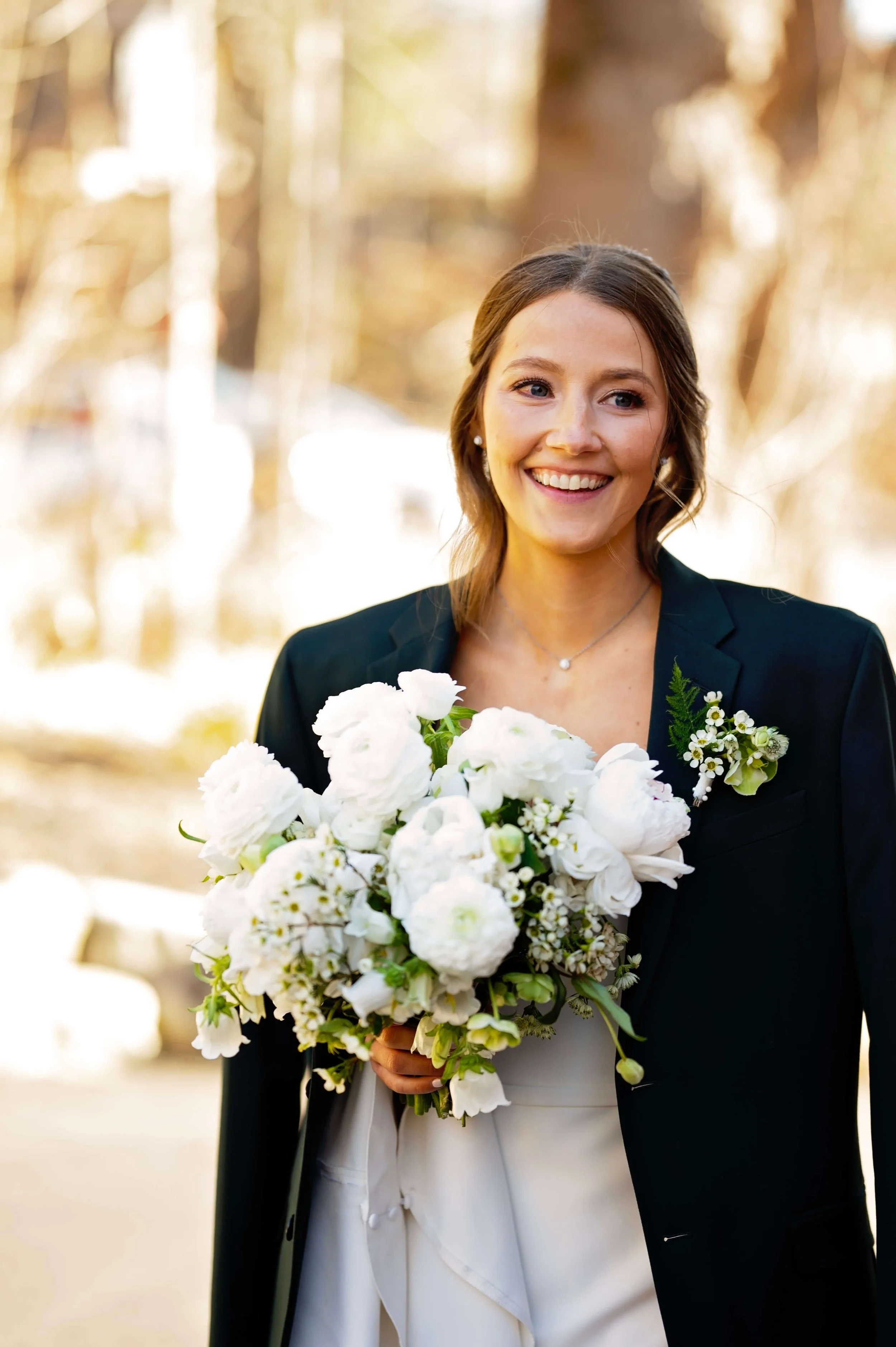Smiling bride in a black blazer holding a lush white bouquet of ranunculus, garden roses, wax flower, and hellebores, Newfound Lake Inn, Bridgewater New Hampshire, Milkweed Floral Co