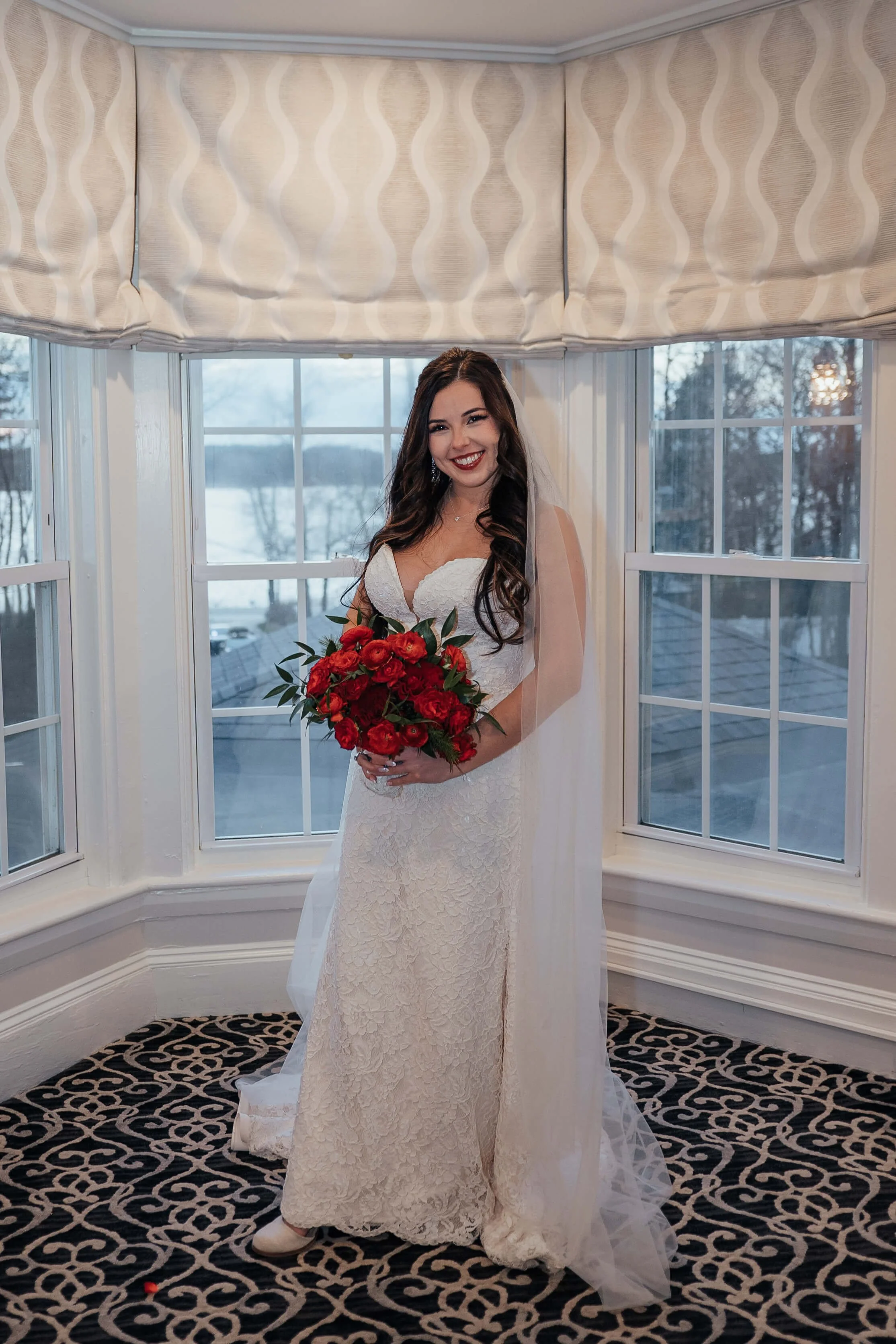Smiling bride in a lace gown holding a lush red peony and garden rose bouquet in a bay window with a snowy winter landscape, New England winter wedding by Milkweed Floral Co