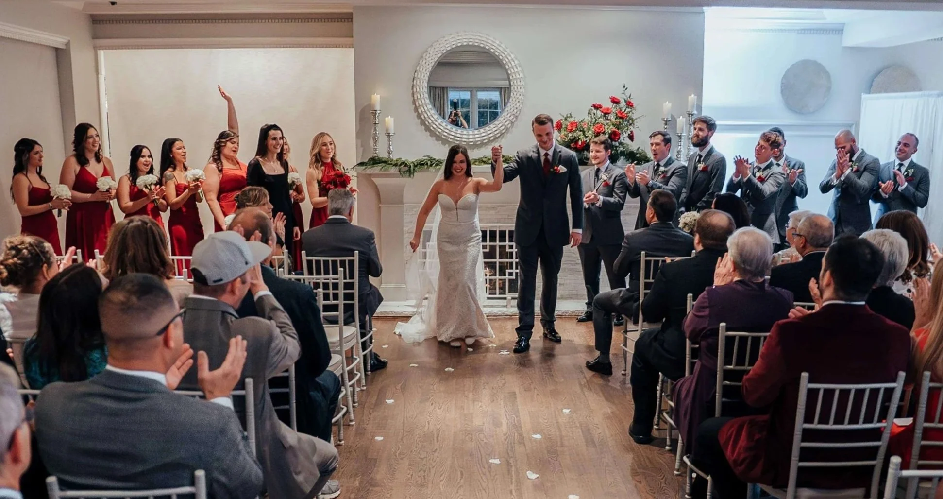 Bride and groom celebrating their recessional with hands raised, bridesmaids in red dresses and red rose fireplace arrangement in the background at Saphire Estate, Sharon Massachusetts wedding florals by Milkweed Floral Co