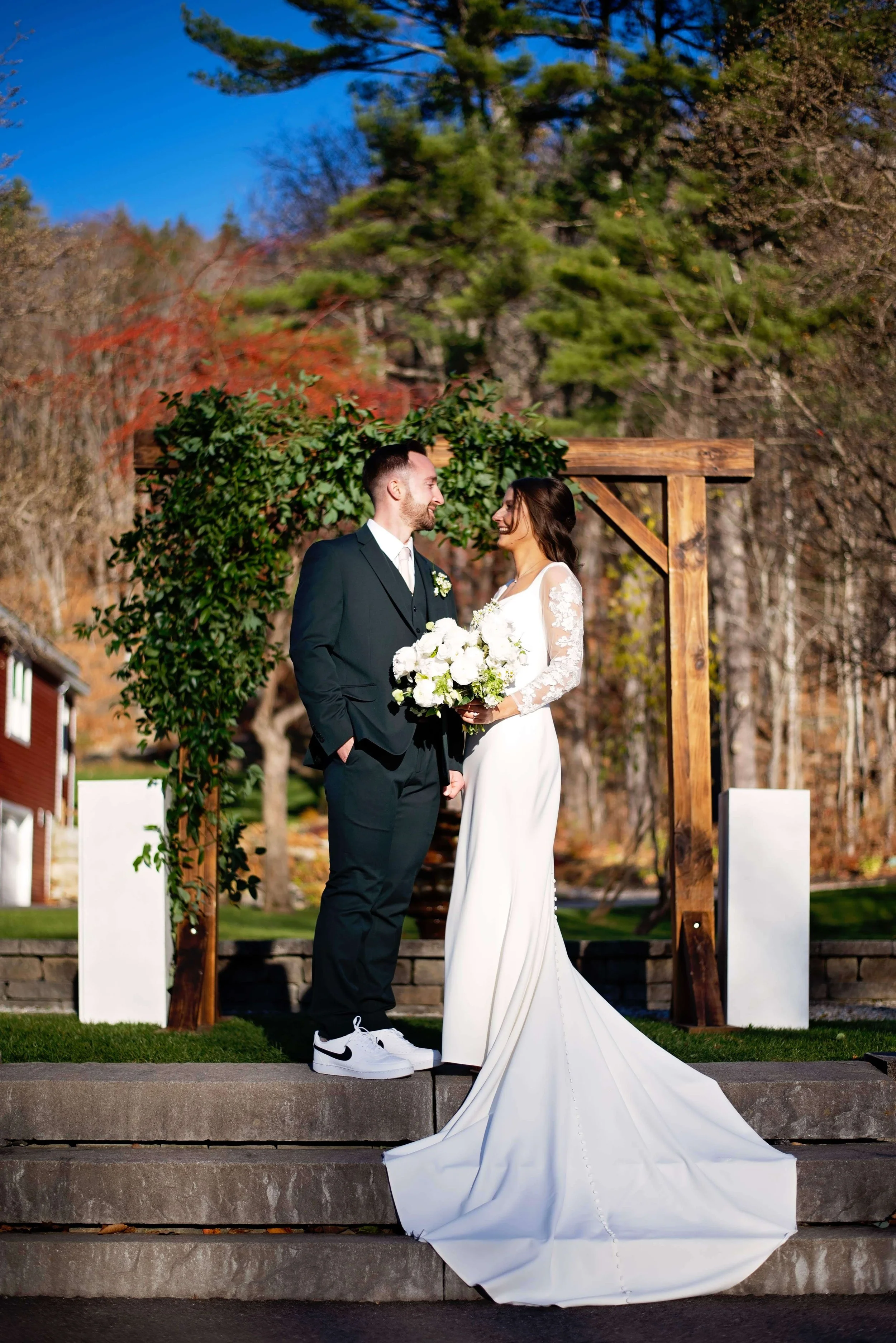 Bride and groom standing before a wood arbor draped with lush greenery, bride holding a white peony and rose bouquet at Newfound Lake Inn, Bridgewater New Hampshire wedding florals by Milkweed Floral Co