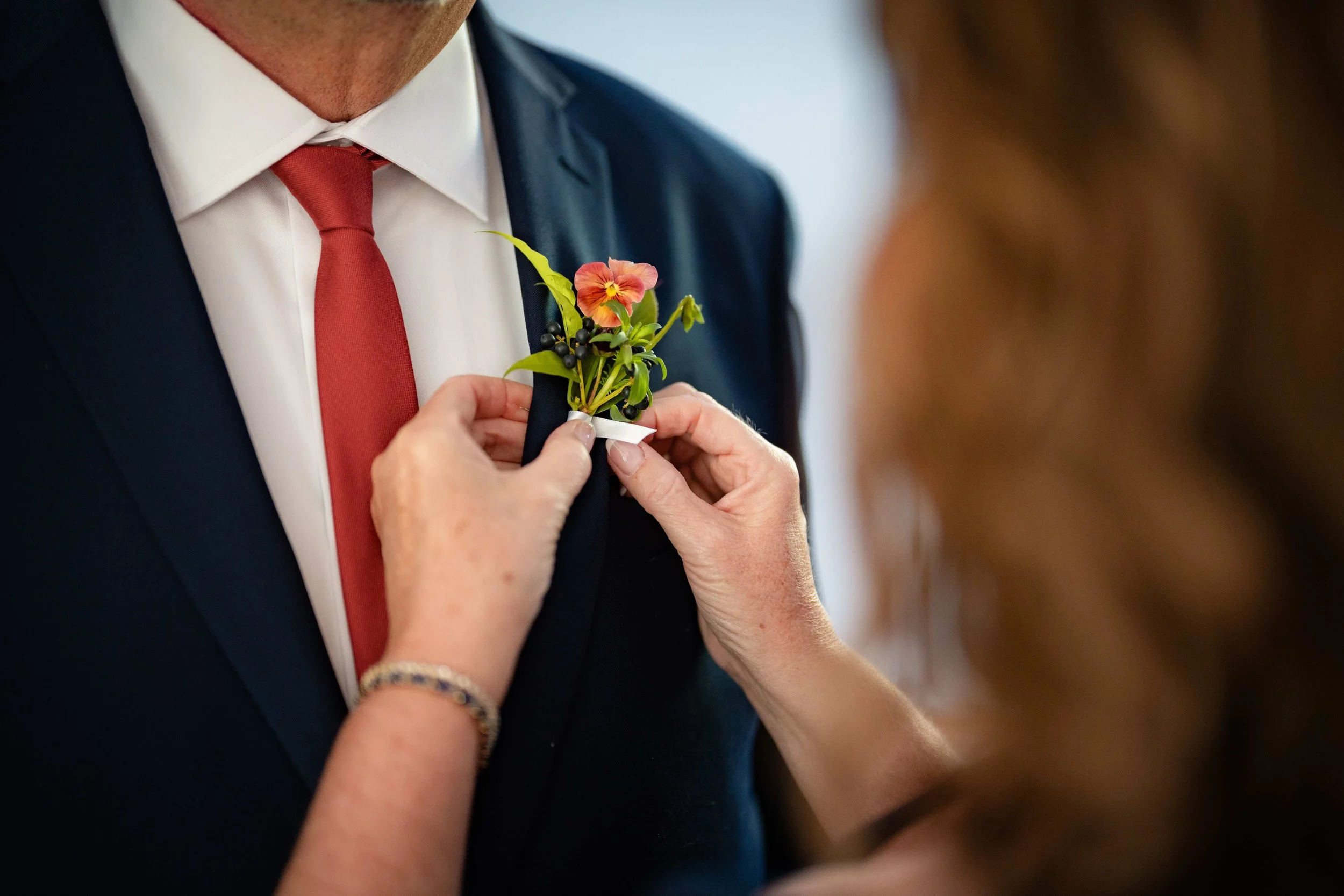 Garden boutonniere with peach pansy and dark berries being pinned to a navy suit, wild-inspired wedding florals by Milkweed Floral Co