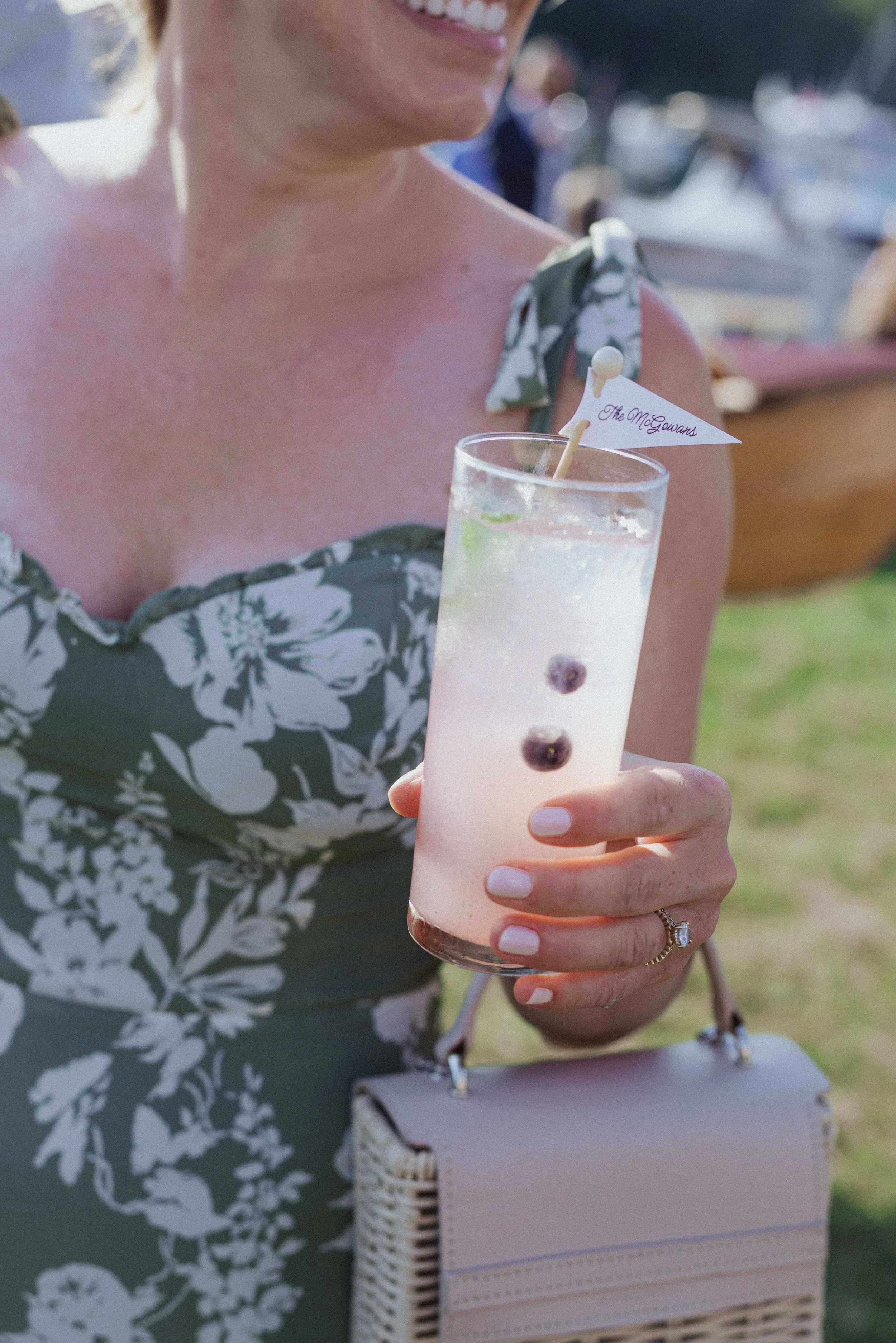 Wedding guest in a floral green dress holding a signature cocktail with a personalized "The McGowans" flag pick at a Nonantum Resort in Kennebunkport