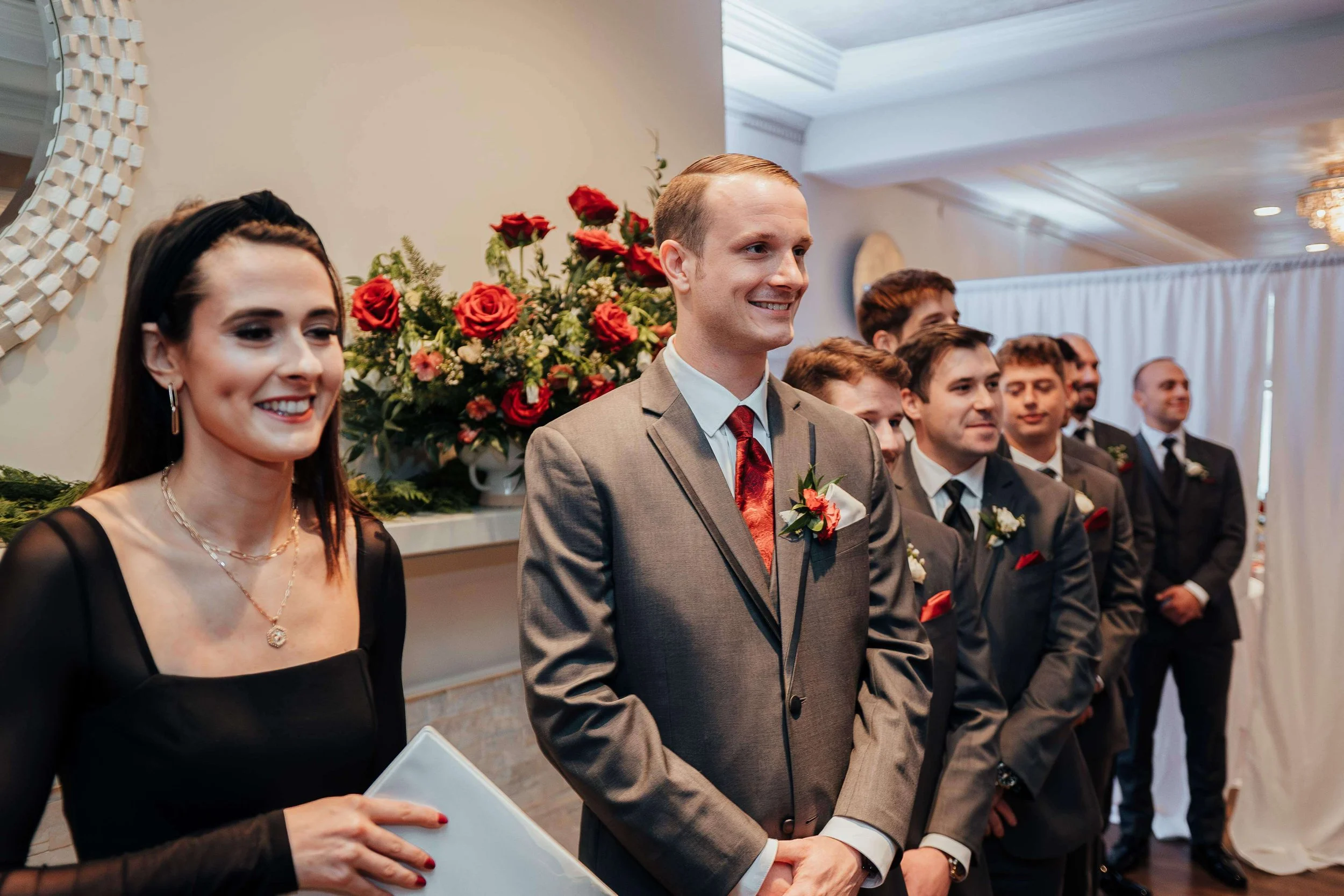 Groom and groomsmen wearing red rose boutonnieres waiting at the altar with a lush red rose ceremony arrangement in the background at Saphire Estate, Sharon Massachusetts wedding florals by Milkweed Floral Co