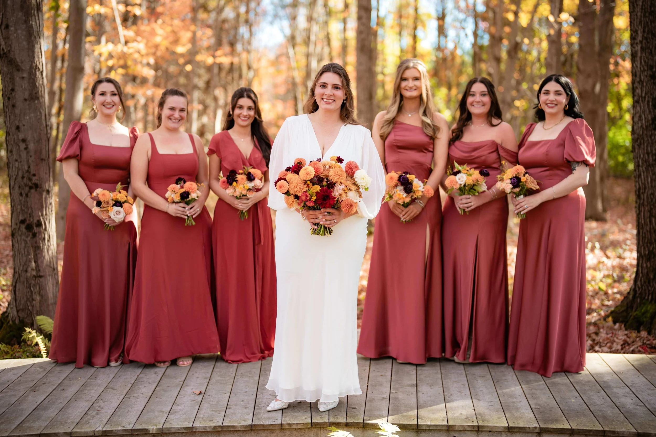 Bride holding bright bouquet of dahlias, standing with bridesmaids in matching berry colored dresses, The Barn at Walnut Hill in North Yarmouth, Maine, fall wedding florals by Milkweed Floral Co