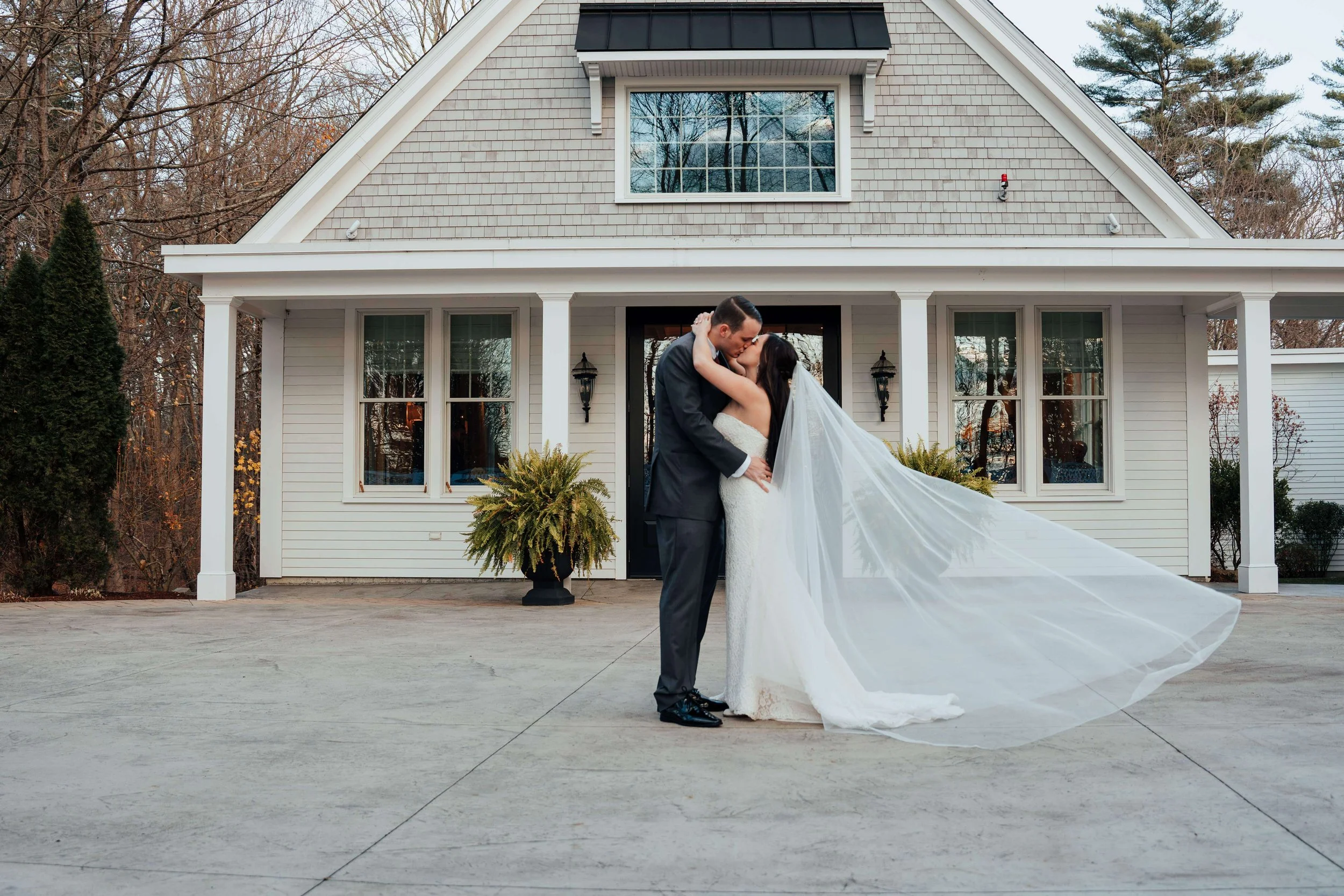 Bride and groom kissing with veil blowing in the wind in front of Saphire Estate, Sharon Massachusetts wedding florals by Milkweed Floral Co