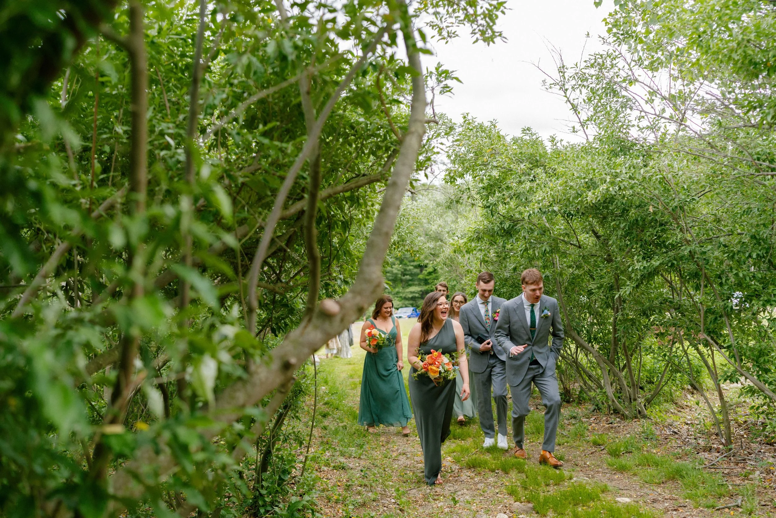 Wedding party walking through a lush green tree-lined path, bridesmaids in sage green dresses holding vibrant orange and yellow wildflower bouquets, Montague Massachusetts spring wedding by Milkweed Floral Co