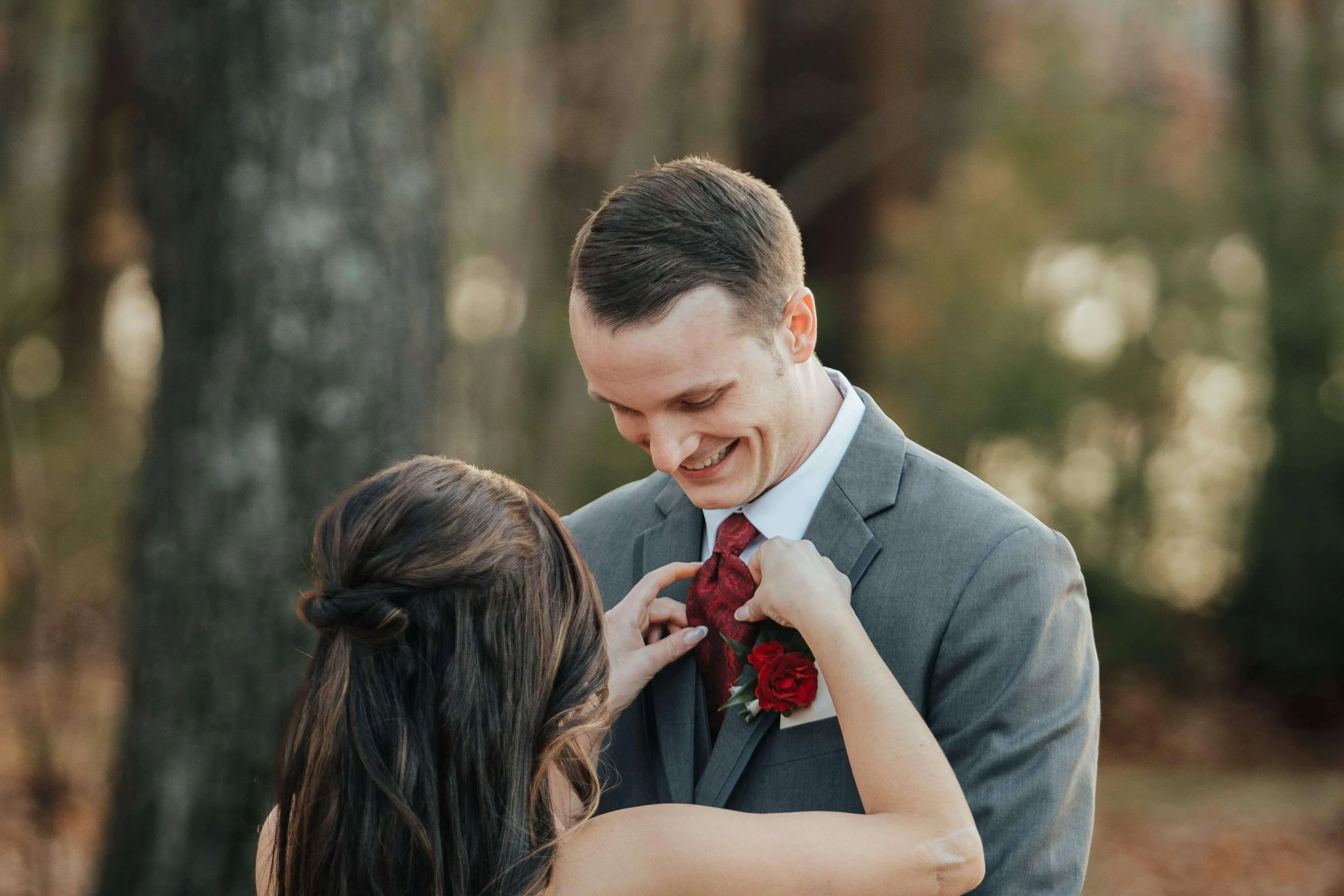 Bride straightening the groom's red tie beside his deep red rose boutonniere in a fall forest, intimate getting ready detail for a New England wedding by Milkweed Floral Co