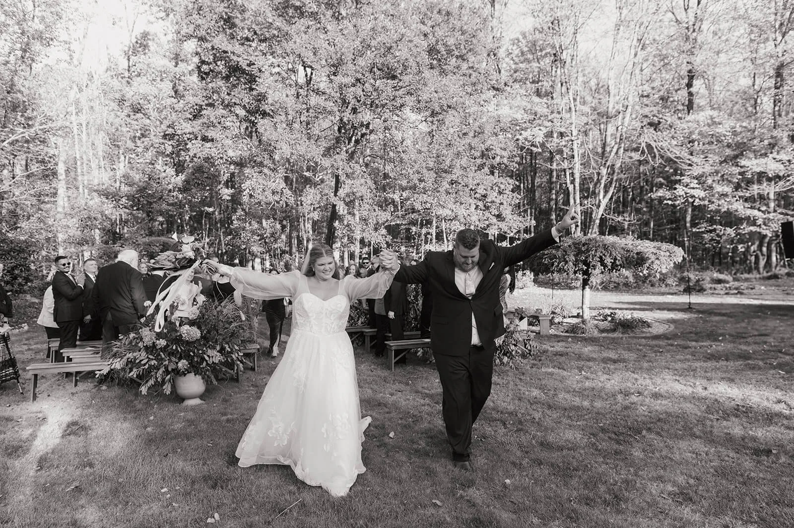 Bride and groom celebrating their recessional with arms raised at an outdoor backyard wedding ceremony, large lush floral urn arrangement in the background, Monson Massachusetts wedding florals by Milkweed Floral Co