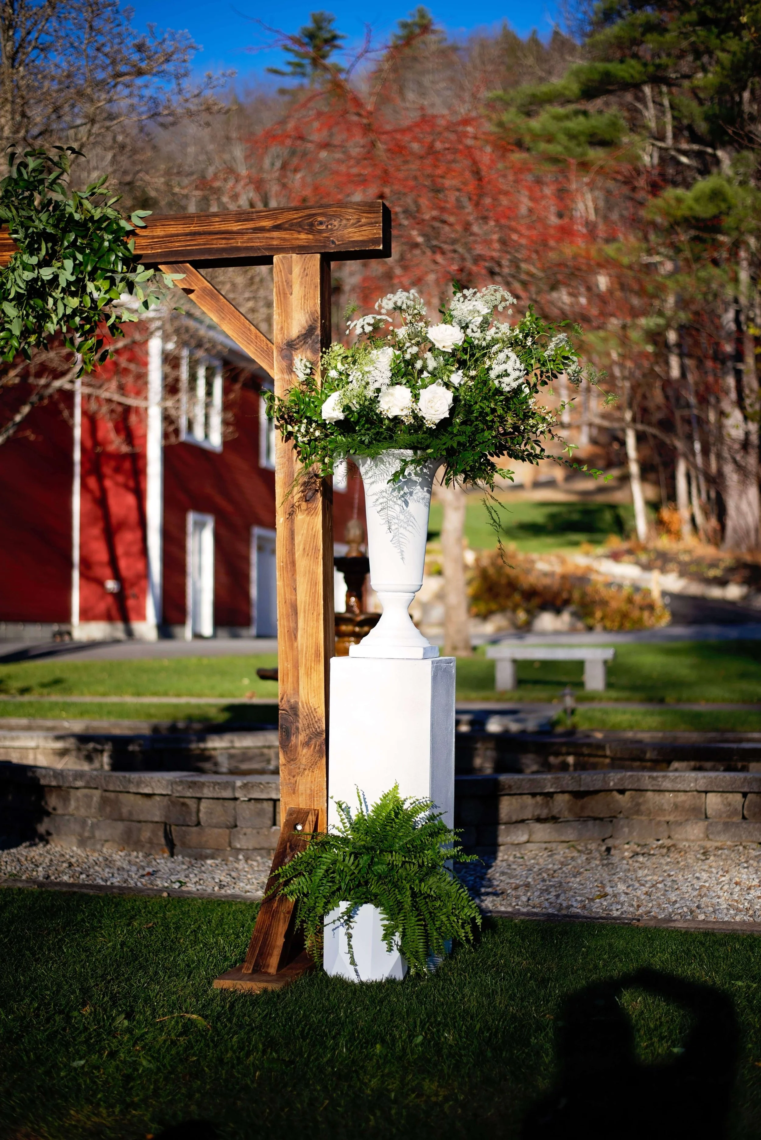 White urn on a pedestal filled with white garden roses, wax flower, and lush greenery beside a wooden ceremony arbor at Newfound Lake Inn, Bridgewater New Hampshire, Milkweed Floral Co