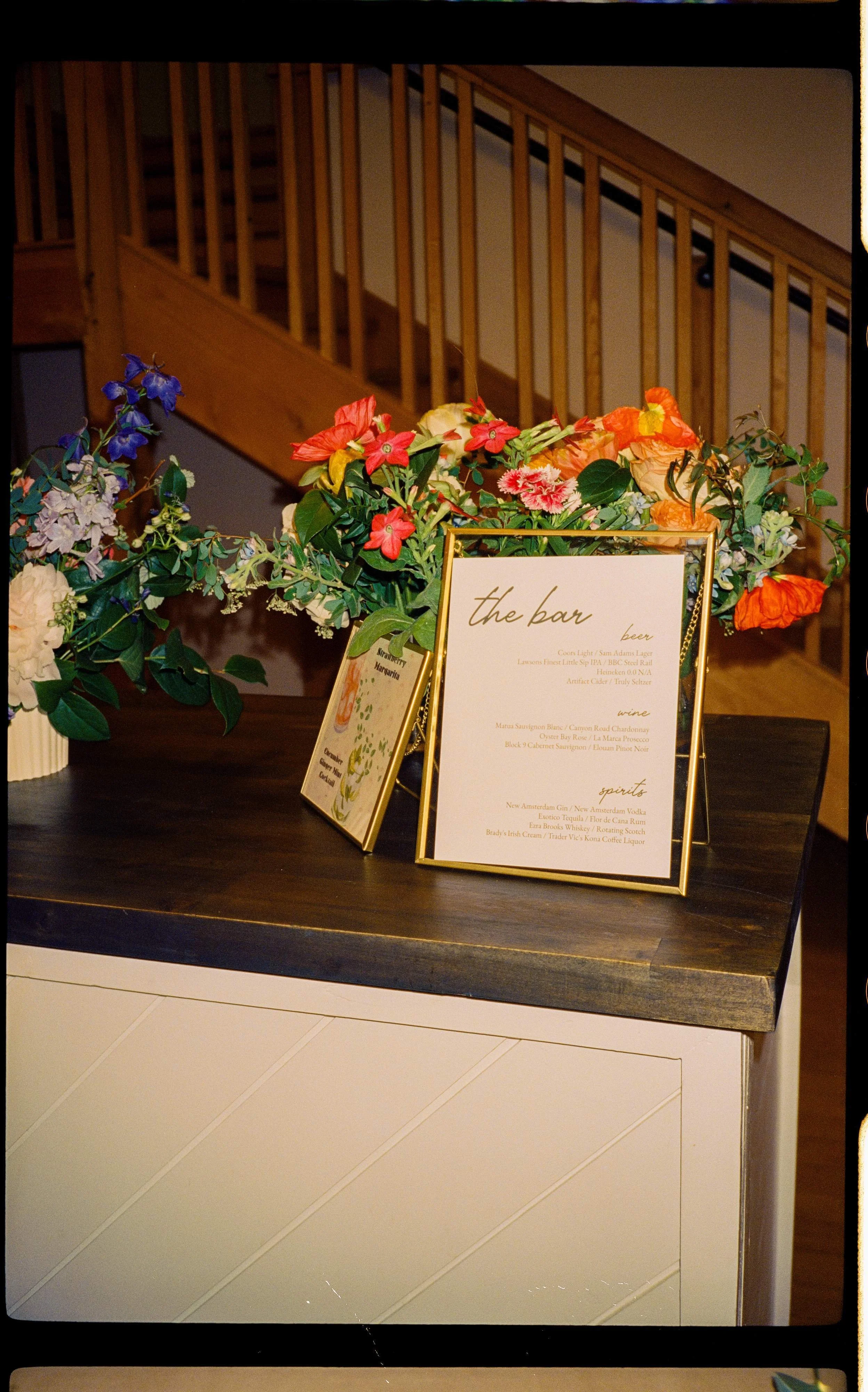 Wedding bar arrangement with orange poppies, red clematis, blue delphinium, and trailing greenery framing a gold-framed bar menu sign, Montague Retreat Massachusetts, Milkweed Floral Co