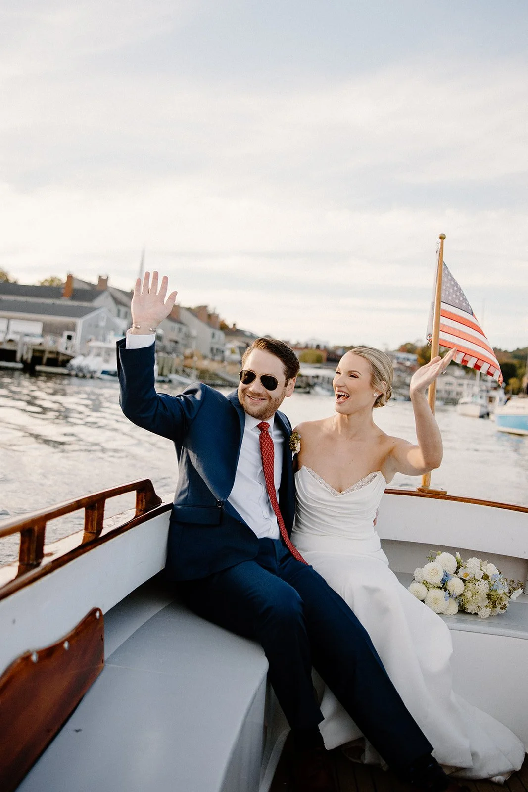 Joyful bride and groom waving from a classic wooden boat in Camden harbor, bride with a white dahlia and blue delphinium bouquet, coastal Maine wedding by Milkweed Floral Co