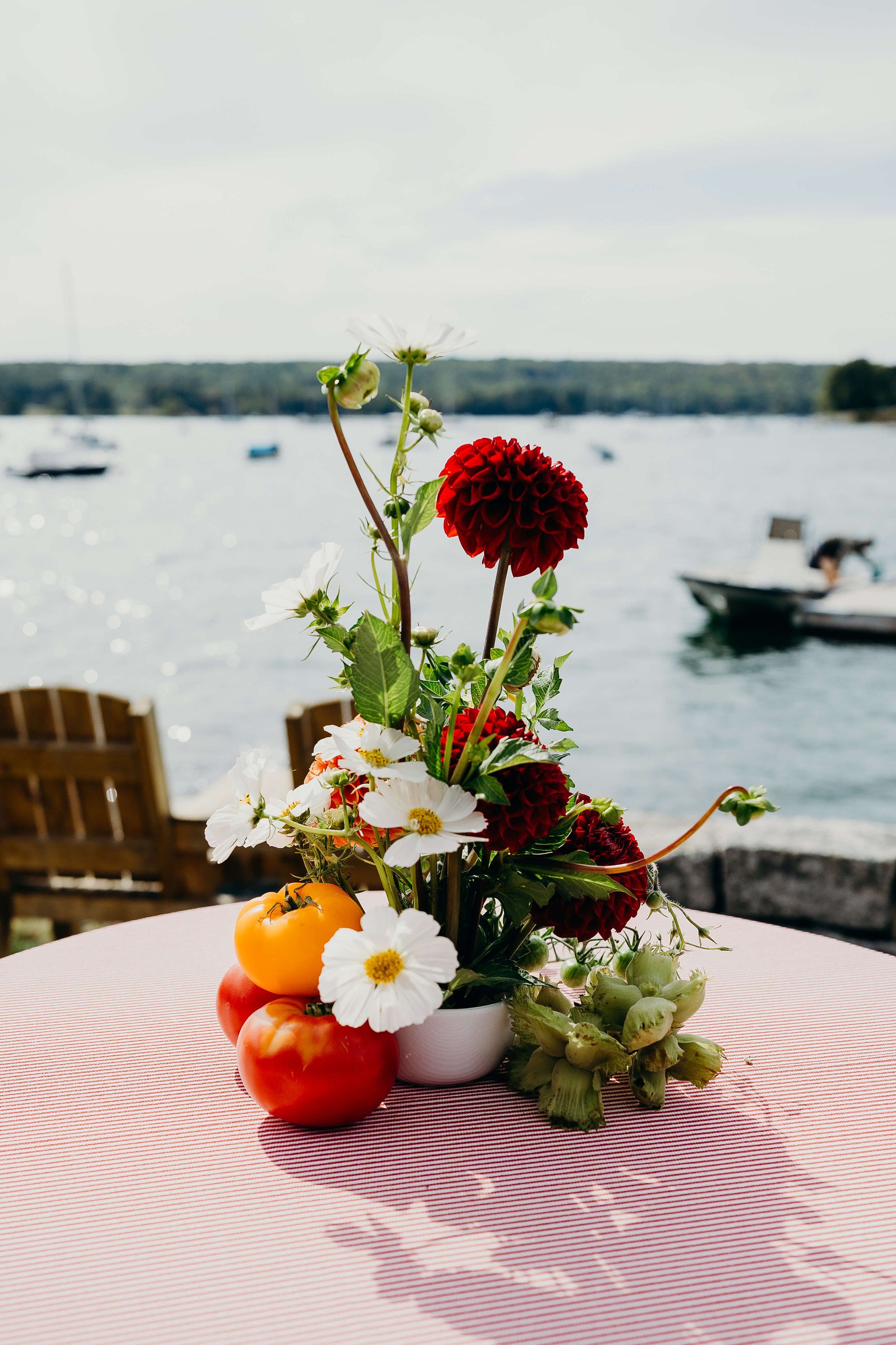 Whimsical coastal wedding centerpiece with deep red dahlias, white cosmos, and heirloom tomatoes on a striped tablecloth overlooking a Maine harbor, Milkweed Floral Co