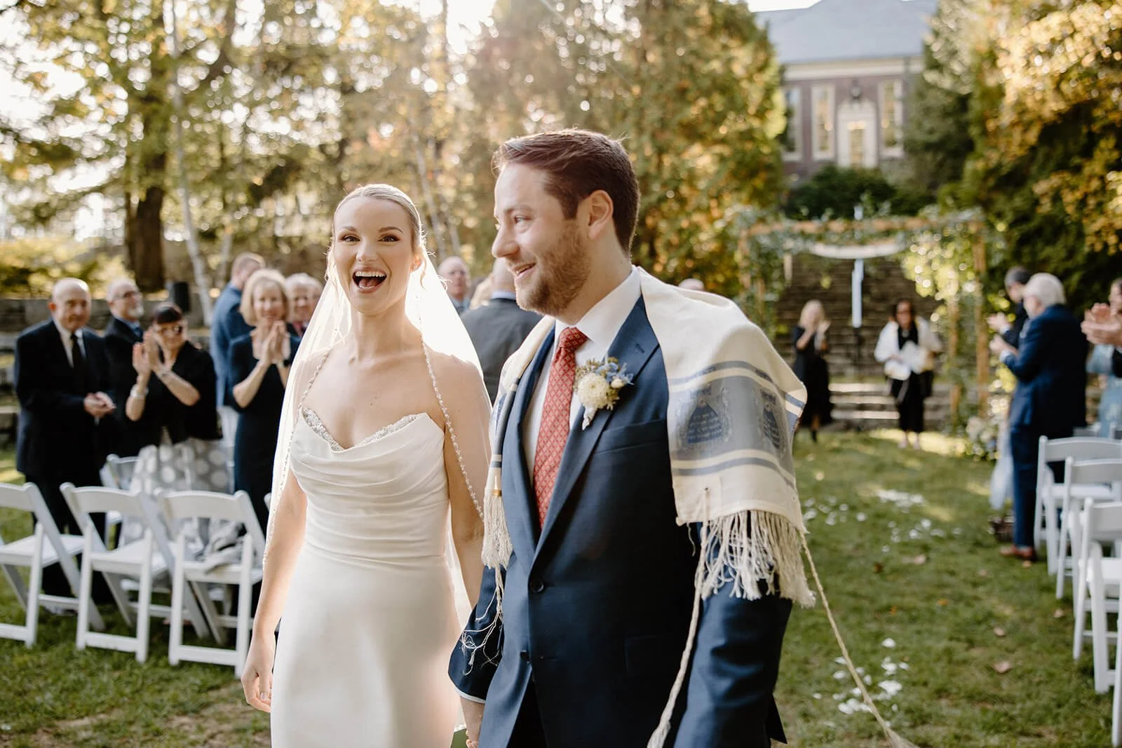 Joyful recessional at Camden Public Library fall wedding with groom wearing a tallit and a floral chuppah in the background, Milkweed Floral Co