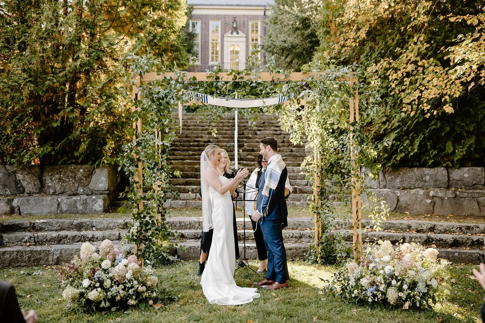 Bride and groom laughing under a greenery-draped chuppah at Camden Public Library with blush and blue hydrangea florals at the base, outdoor fall wedding by Milkweed Floral Co