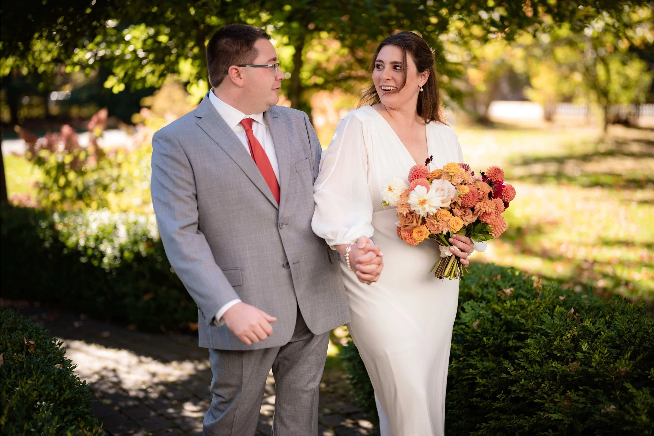 Bride and groom on a sunlit path, bride holding a lush fall bouquet of peach and coral dahlias and garden roses, Milkweed Floral Co