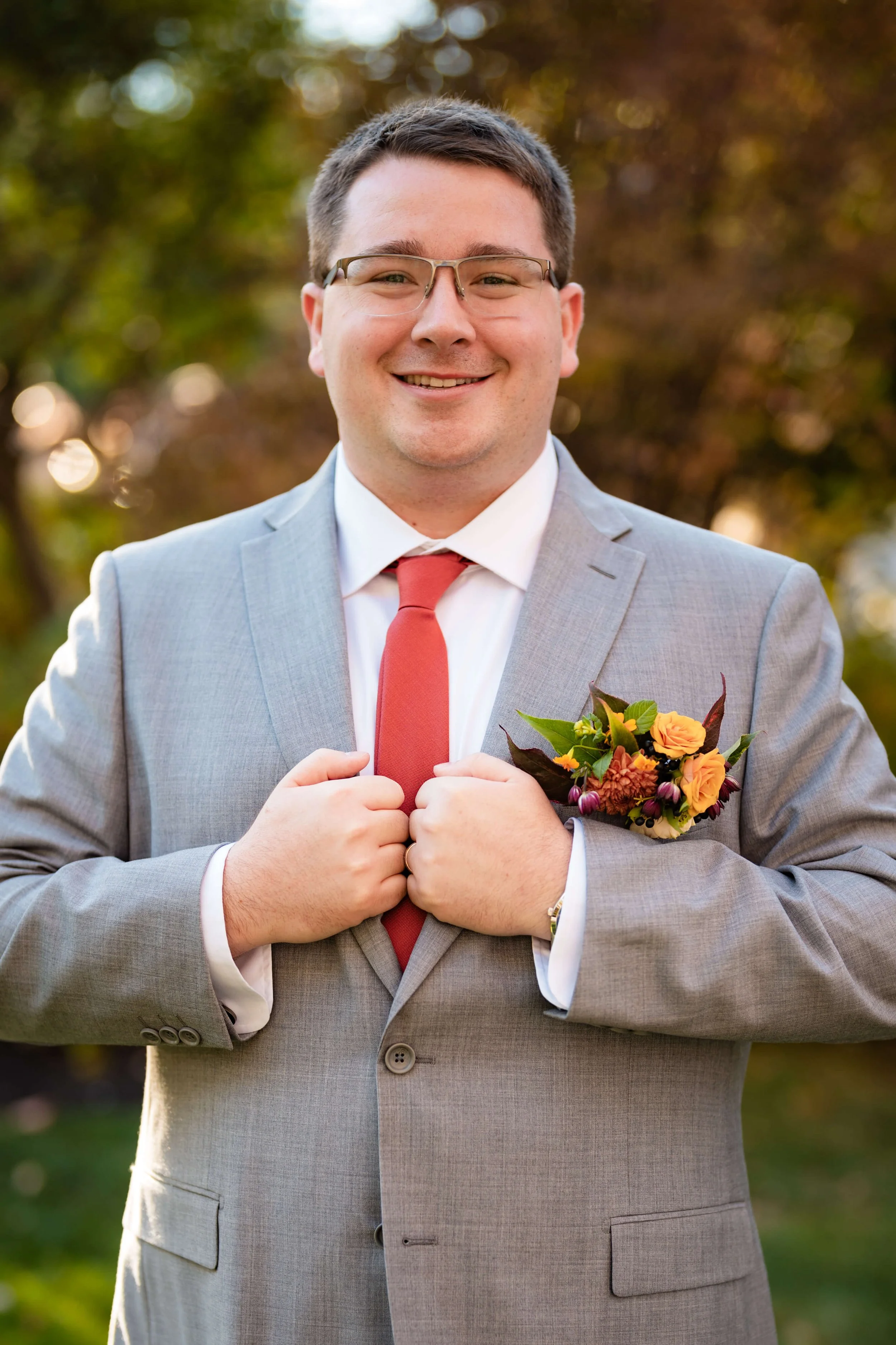 Smiling groom in gray suit with red tie and autumn colored boutonniere,outdoor fall Maine wedding, florals by Milkweed Floral Co