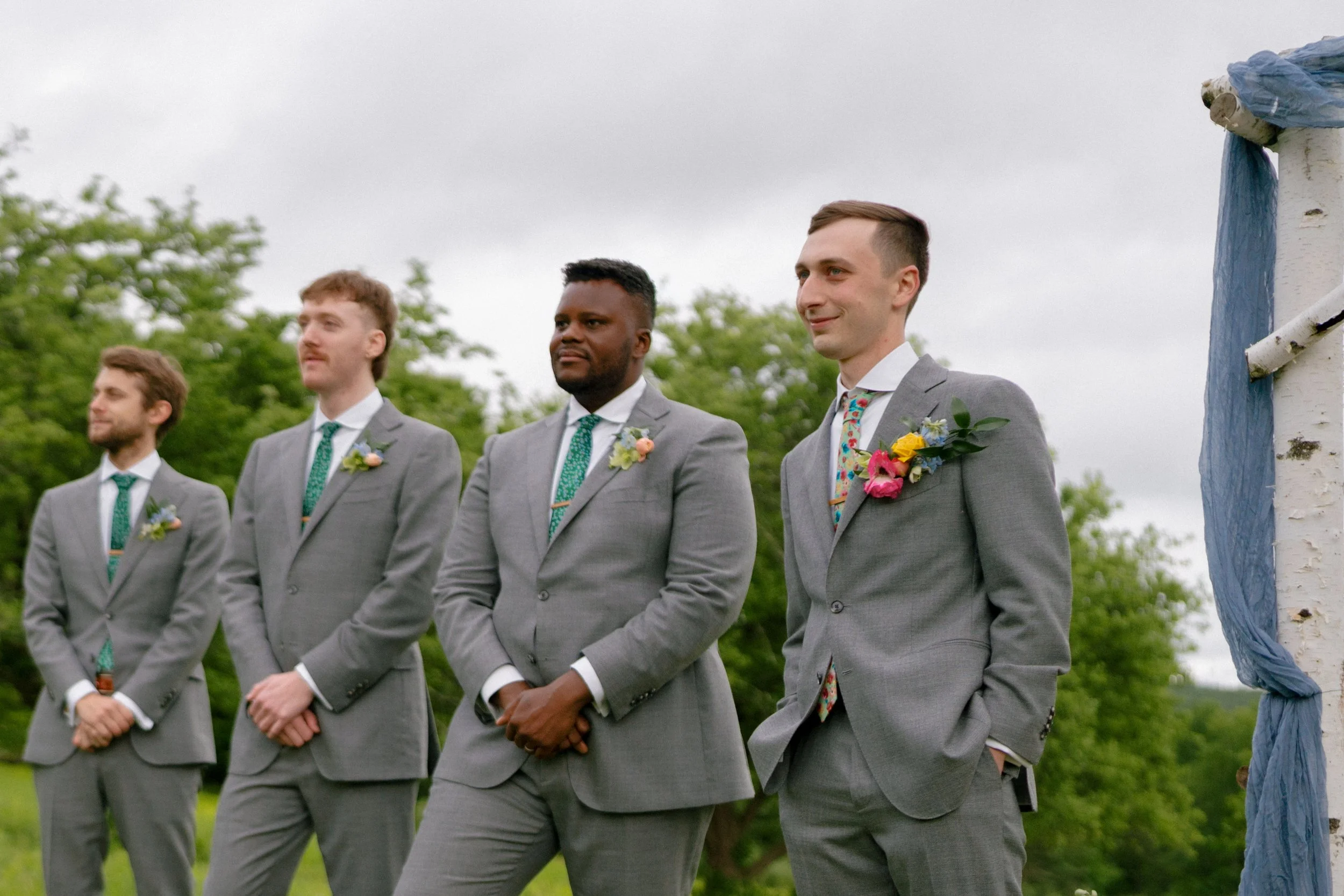 Groom and groomsmen in grey suits with colorful wildflower boutonnieres of poppies, ranunculus, and delphinium at an outdoor spring wedding, Montague Massachusetts, Milkweed Floral Co