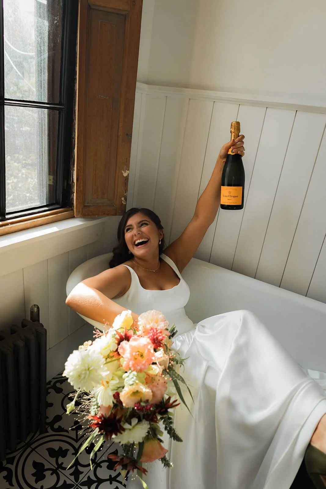 Joyful bride in a bathtub holding a champagne bottle and a lush peach, cream, and burgundy bridal bouquet of dahlias and ranunculus, New England wedding by Milkweed Floral Co