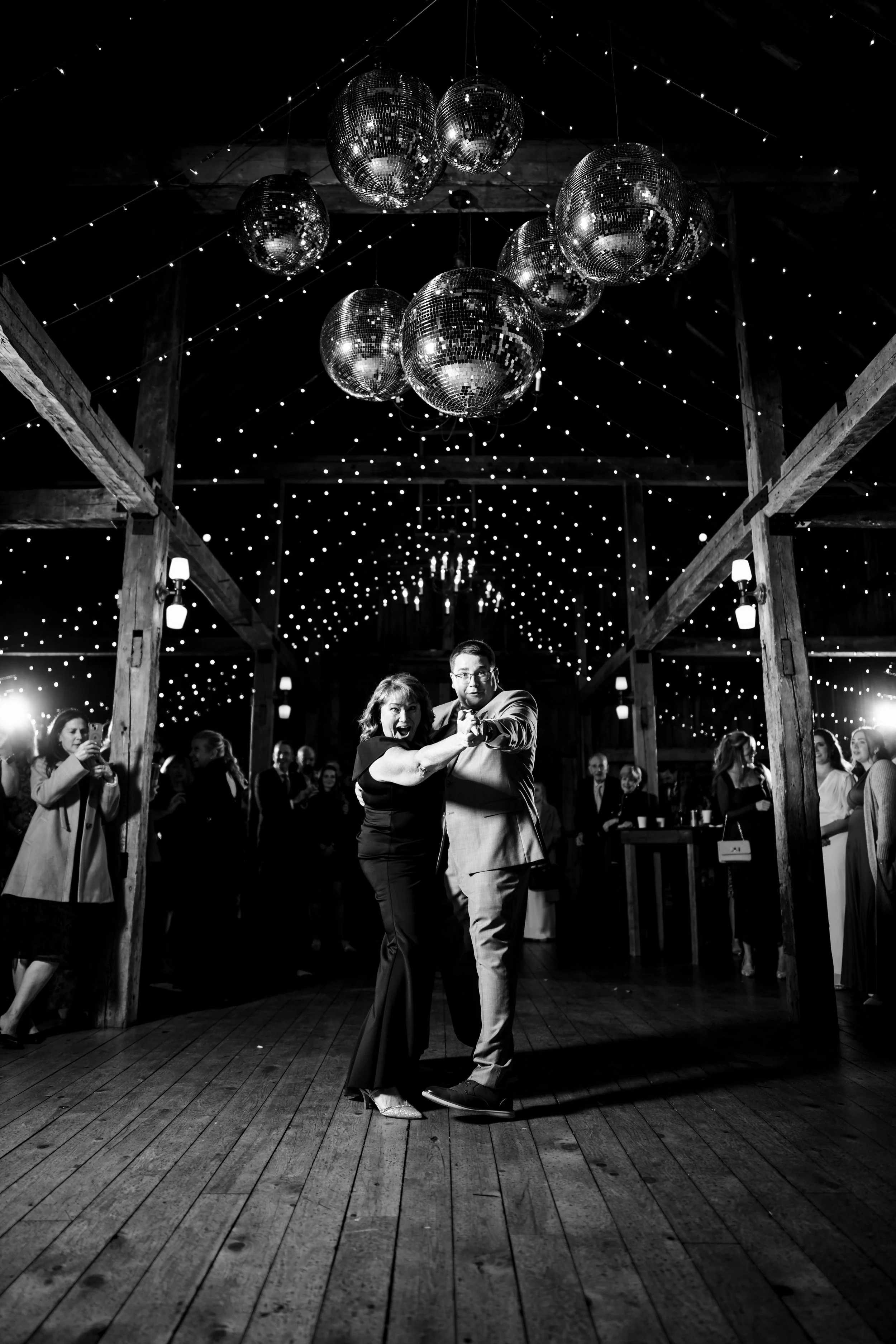 Groom and mother-of-the-groom dancing with arms pointed toward the camera under cluster of disco balls hanging from ceiling and string lights, North Yarmouth, Maine, Milkweed Floral Co