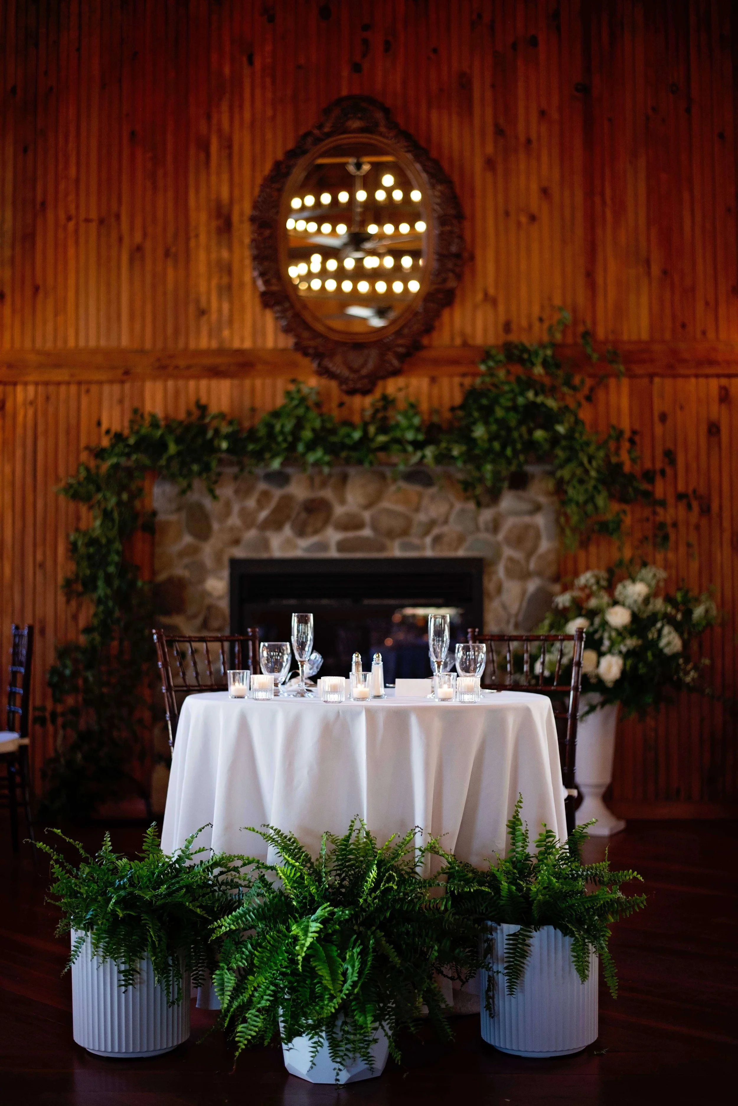 Sweetheart table framed by potted ferns and a greenery-draped stone fireplace at Newfound Lake Inn, Bridgewater New Hampshire wedding florals by Milkweed Floral Co