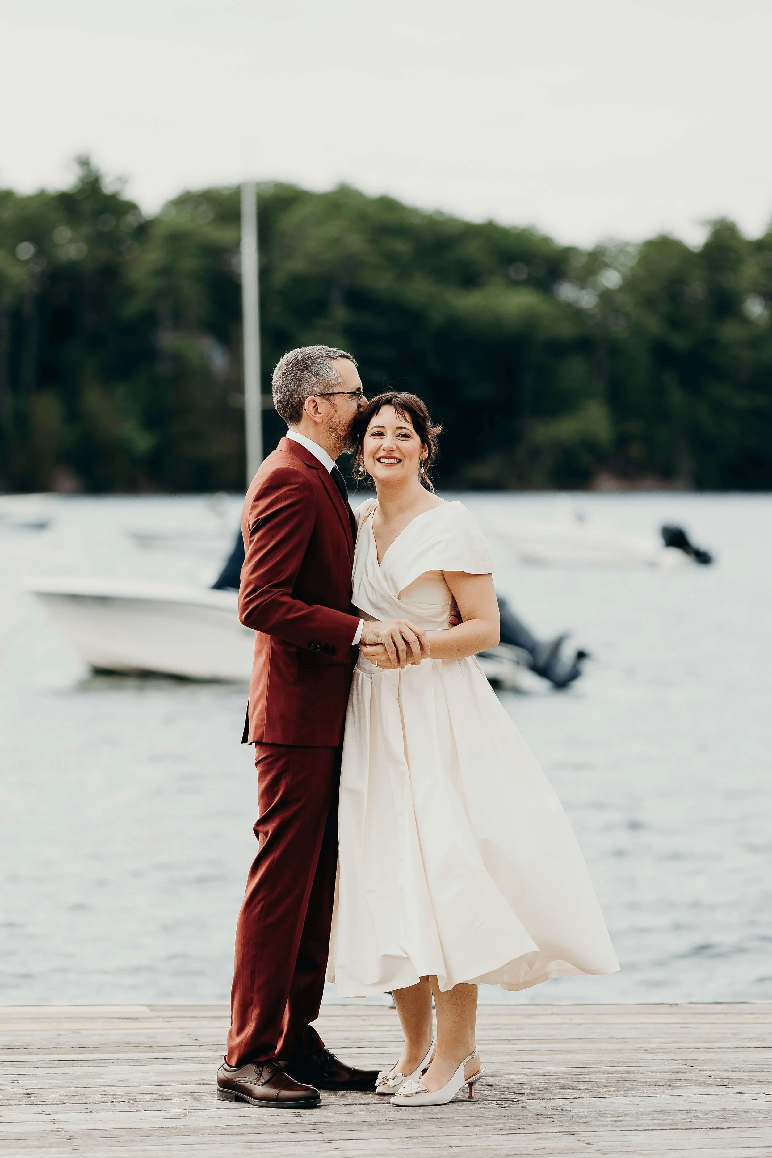 Bride and groom embracing on a coastal Maine dock with sailboats in the background, groom in a burgundy suit, coastal wedding florals by Milkweed Floral Co