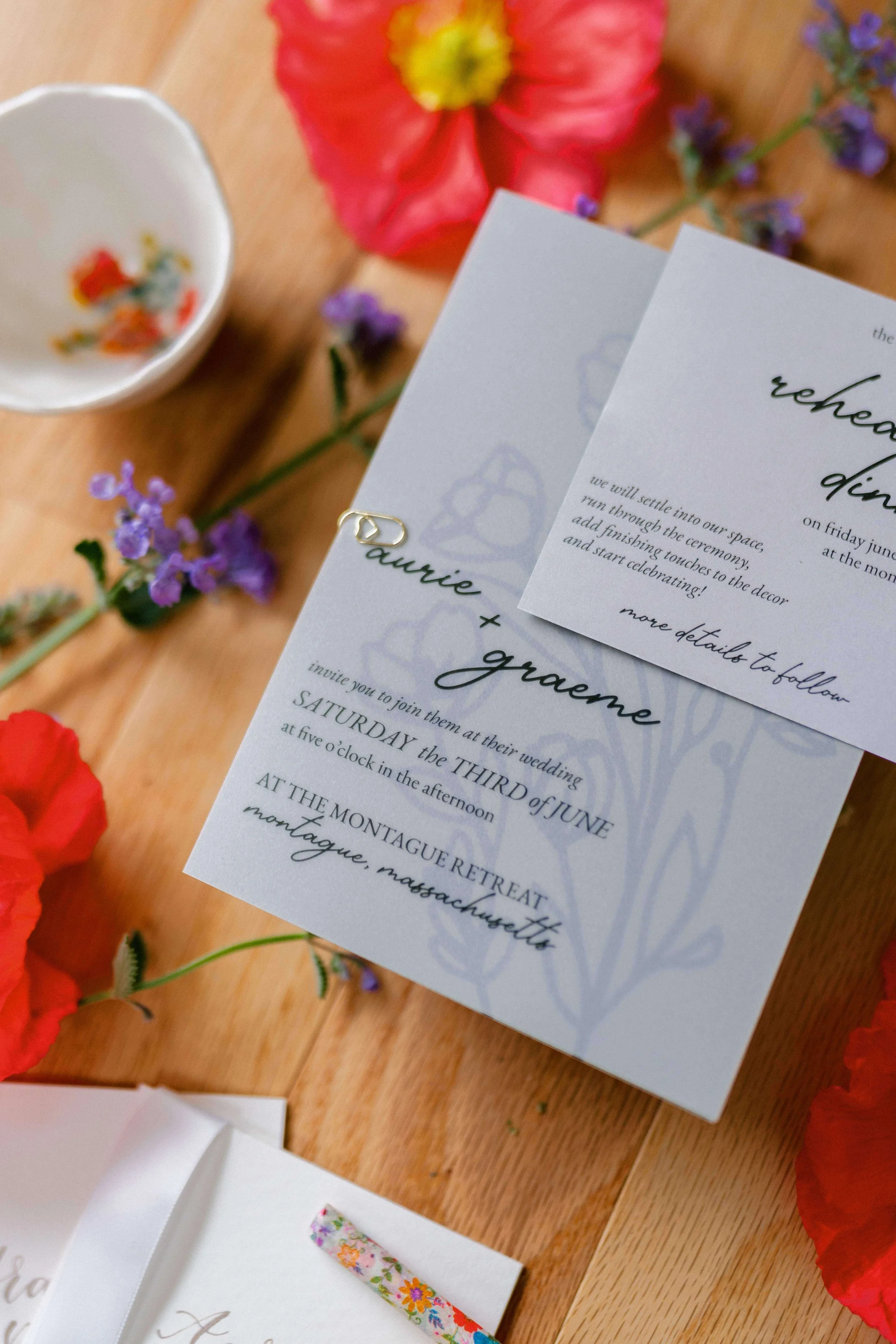 Spring wedding invitation suite flatlay with dusty blue stationery surrounded by red poppies and purple wildflowers, Montague Retreat Massachusetts wedding by Milkweed Floral Co