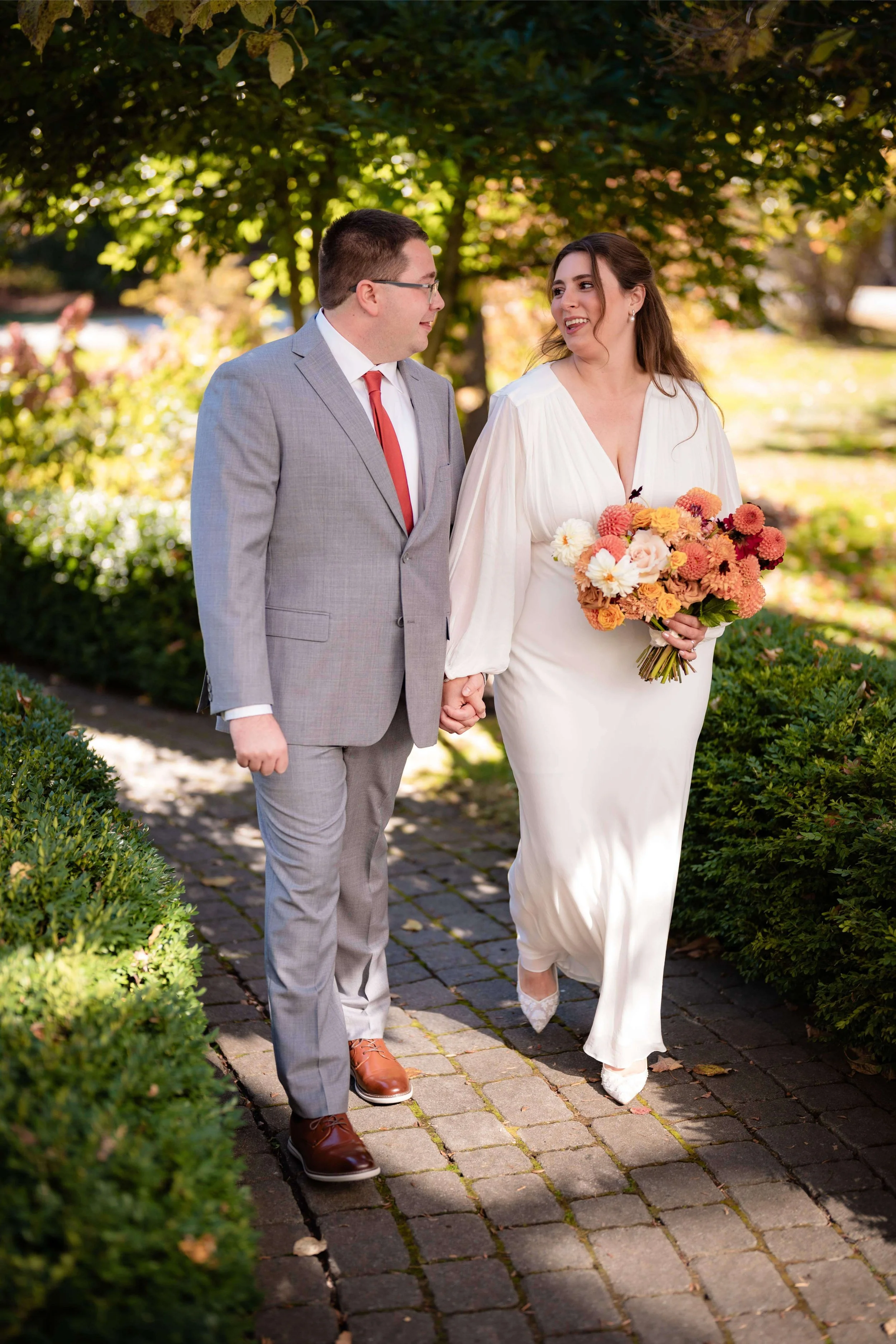 Bride and groom holding hands and smiling on sunlit garden path at the Barn at Walnut Hill with blush and orange dahlia bouquet, outdoor fall wedding by Milkweed Floral Co