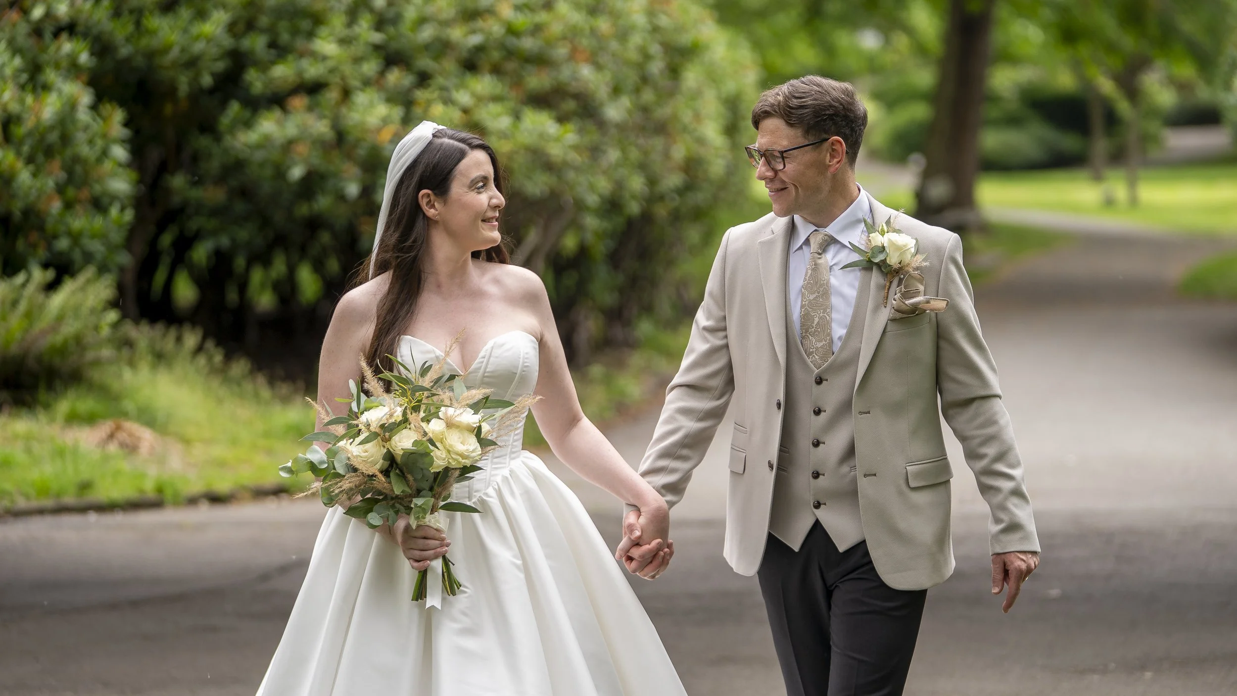 A bride and groom walking hand in hand outdoors, smiling at each other. The bride is in a white wedding gown holding a bouquet, and the groom is in a light-colored suit with a boutonniere.