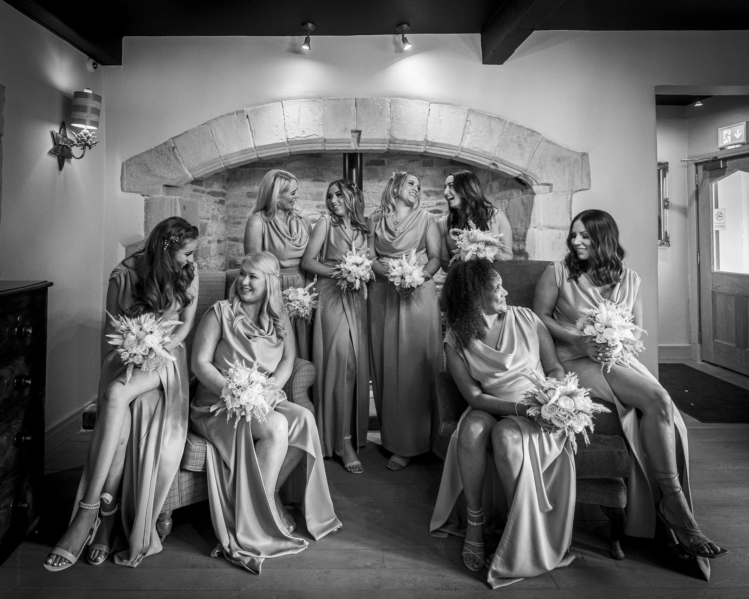 A group of women in satin dresses holding bouquets, sitting and standing in a cozy room with a stone fireplace backdrop, smiling and engaging with each other.