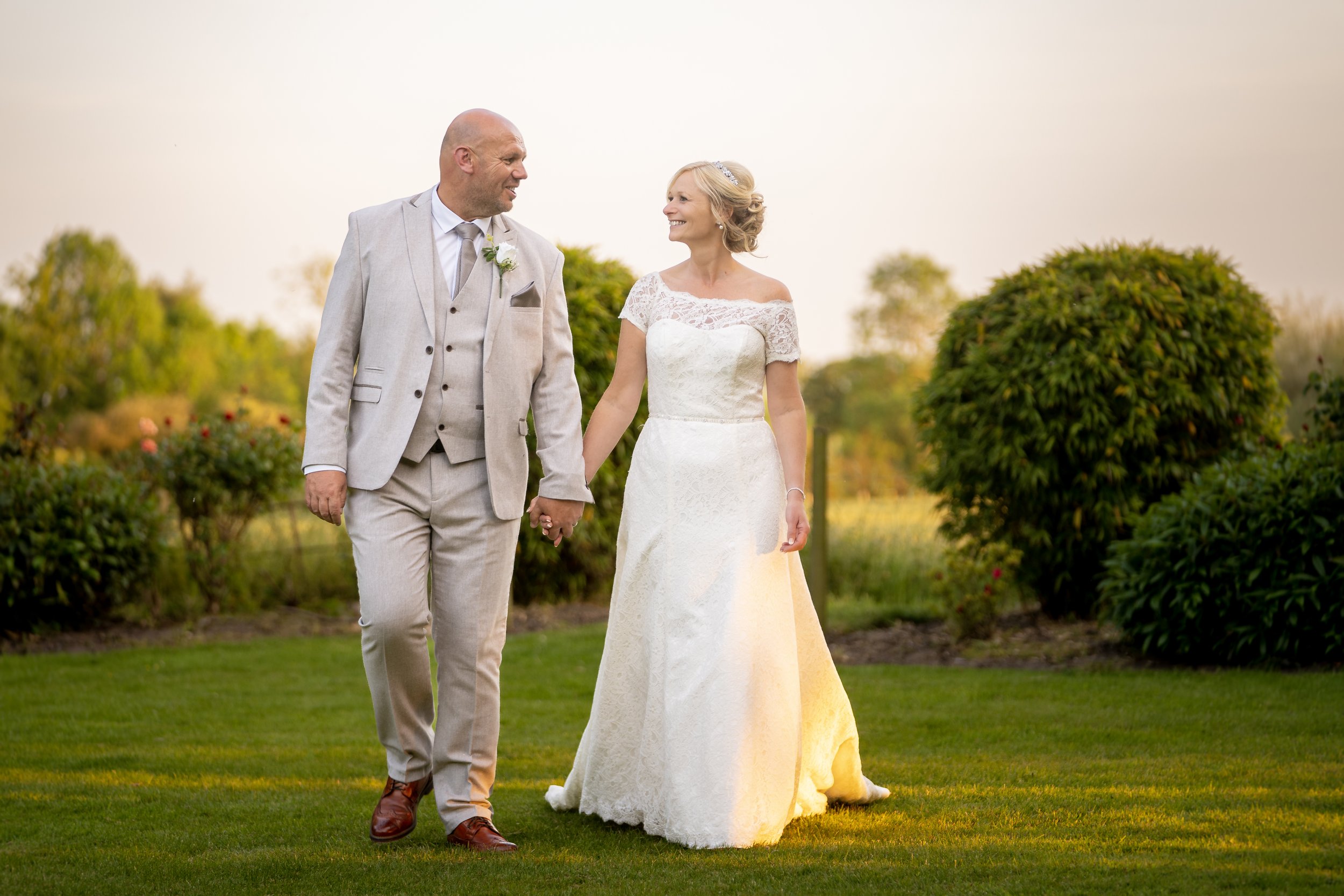 A bride and groom holding hands and walking on a lush green lawn during sunset, surrounded by bushes and trees.