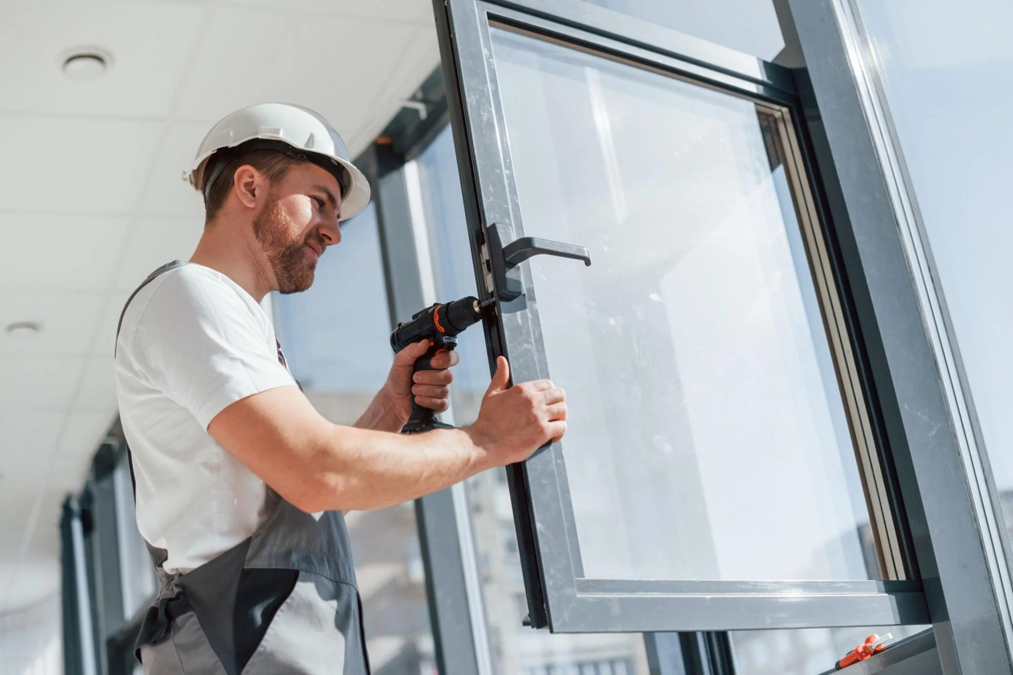 Man wearing a white hard hat and gray apron installing or repairing a window with a cordless drill.