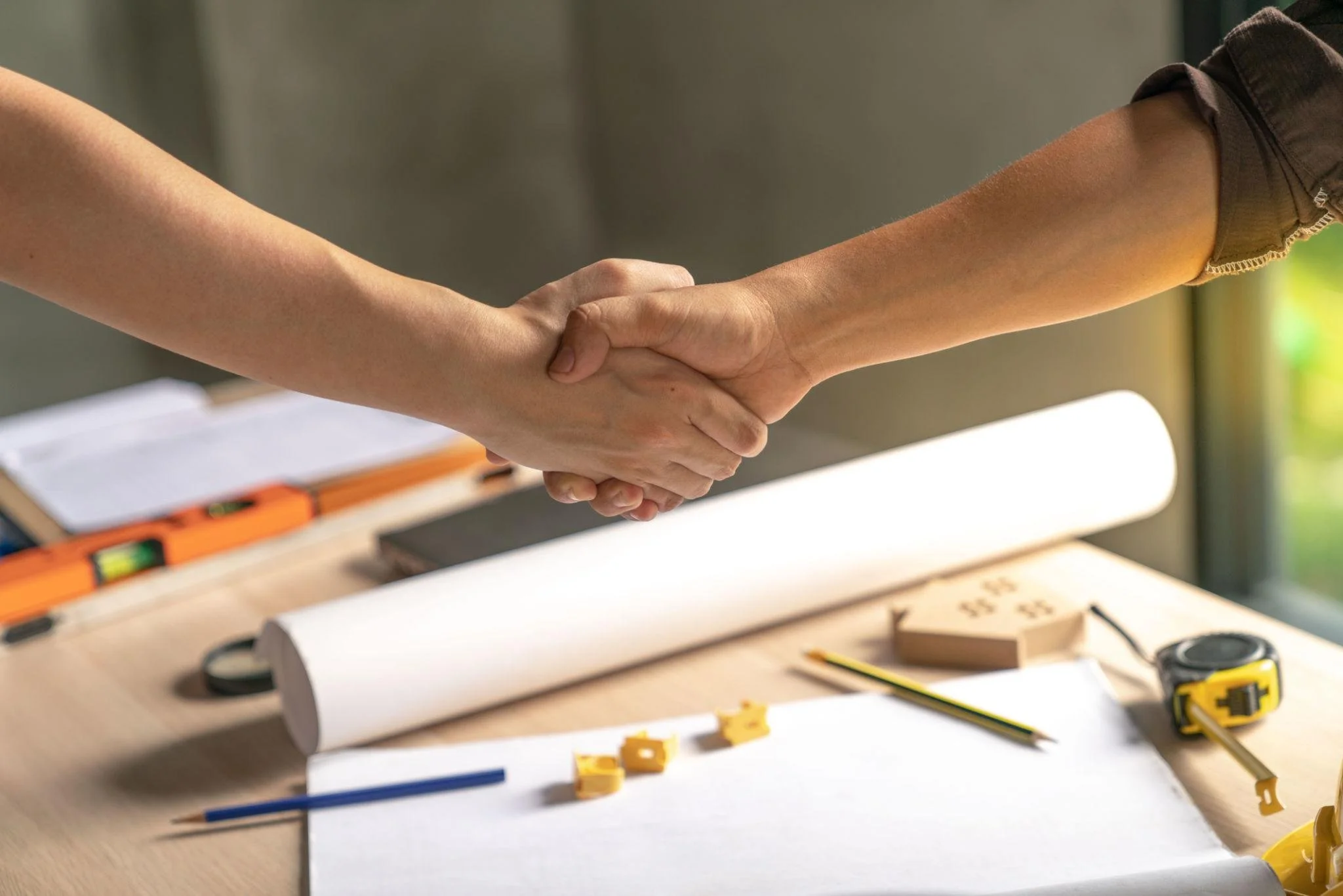 Two people shaking hands over a desk with various tools and papers, possibly in a workspace or drafting area.