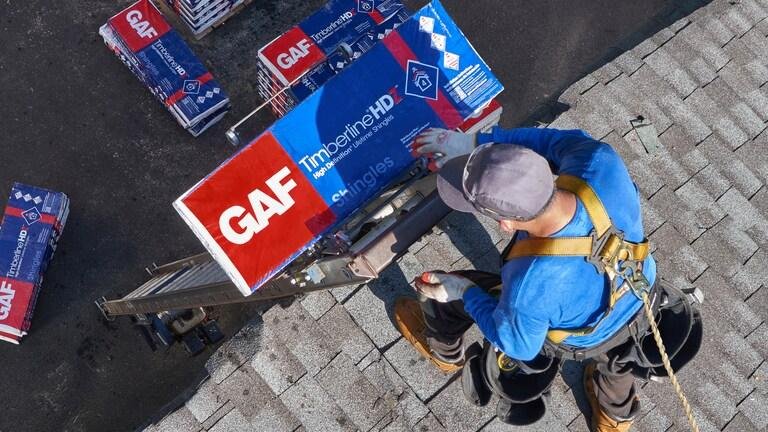 A worker on a roof installing or repairing signage for GAF Timberline HD shingles, with multiple shingles pallets nearby.