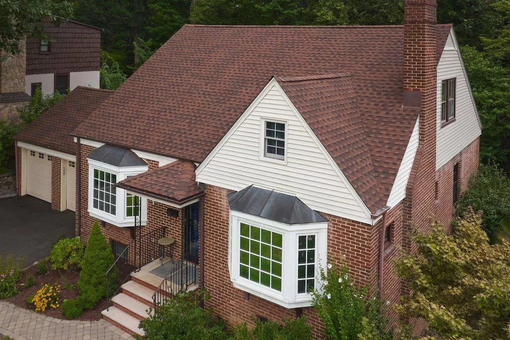 An aerial view of a brick house with a steep gabled roof, white siding, large bay windows, a chimney, and a two-car garage, surrounded by greenery.