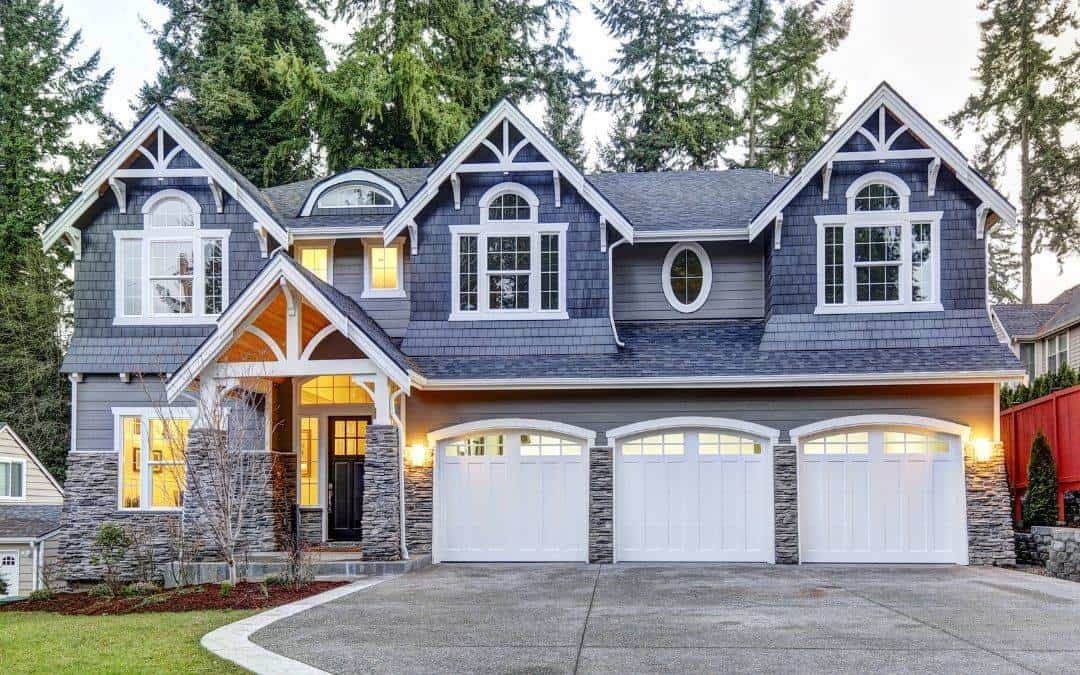 A large two-story house with dark blue siding, white trim, and three garage doors. The house features multiple gables, large windows, and a stone front porch with a black door. The yard has a curved driveway and some landscaping.