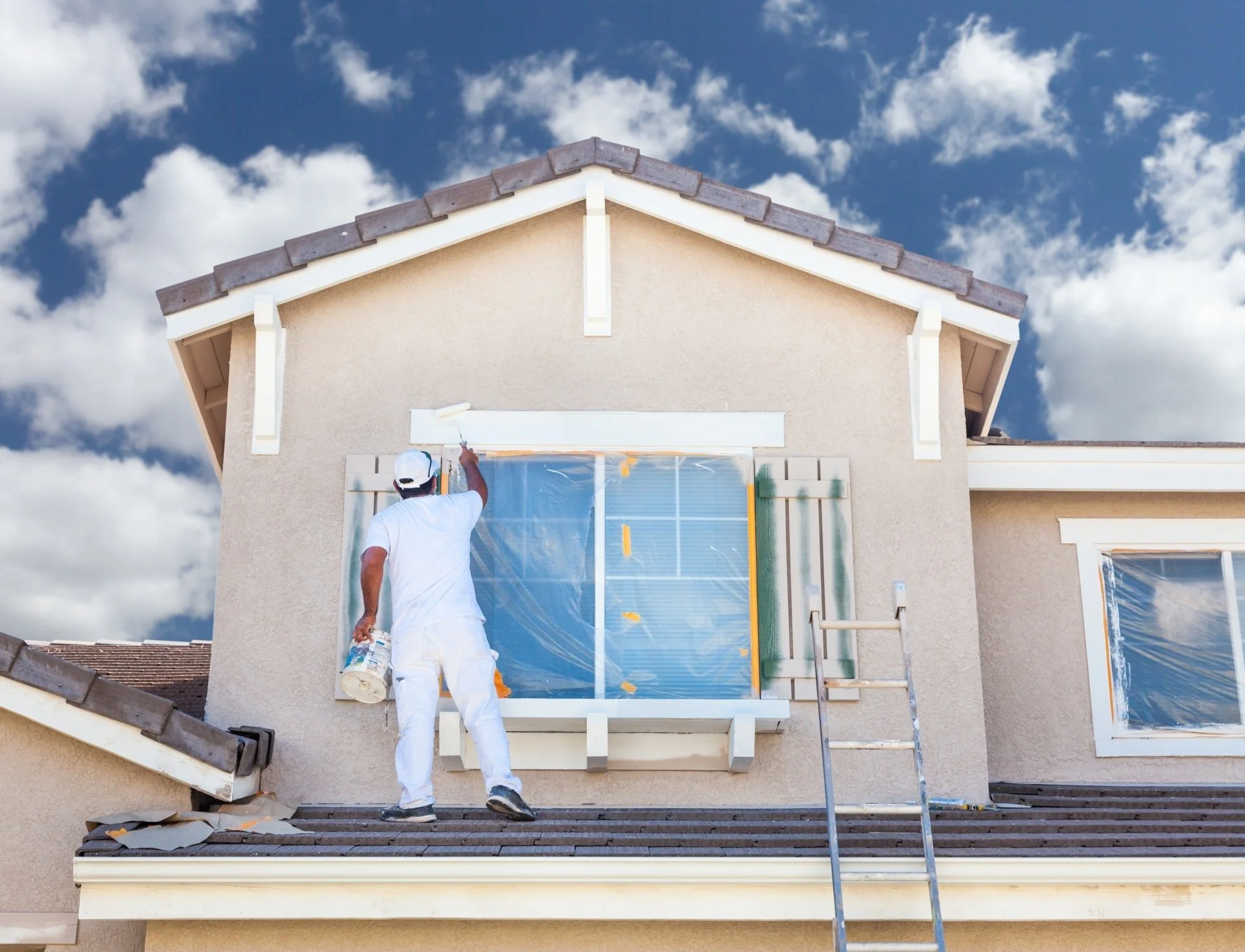 A person in white overalls and a cap painting the trim of a house's second story window with white paint. The house has tan stucco walls and a brown shingled roof. A ladder is positioned against the house, and the sky is partly cloudy.