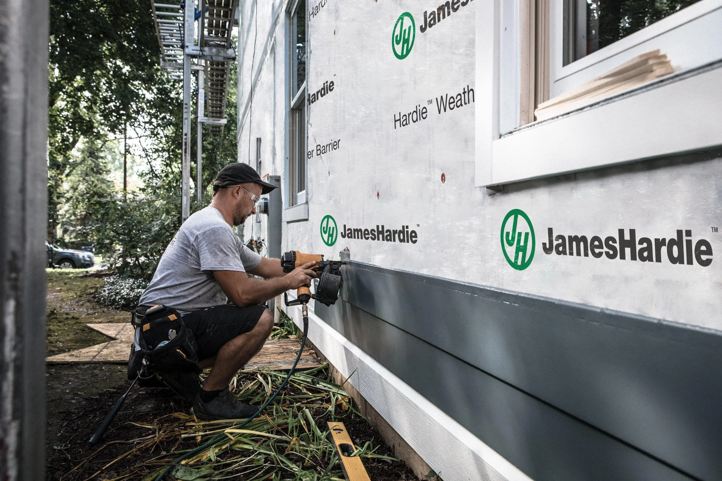 A man installing exterior siding on a building with JamesHardie branding, using a power tool, squatting on the ground, surrounded by trees and construction materials.